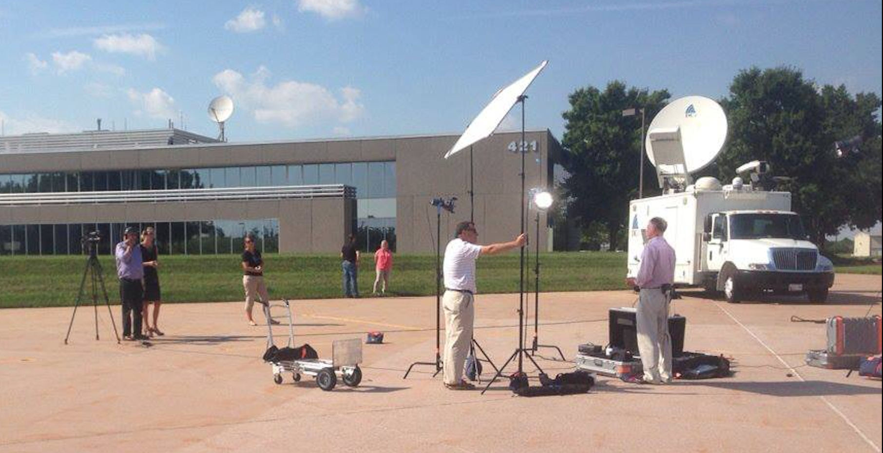 A crew of people are operating television equipment on an airplane ramp. There is a satellite uplink truck with its dish up. A corporate office building is visible in the background.