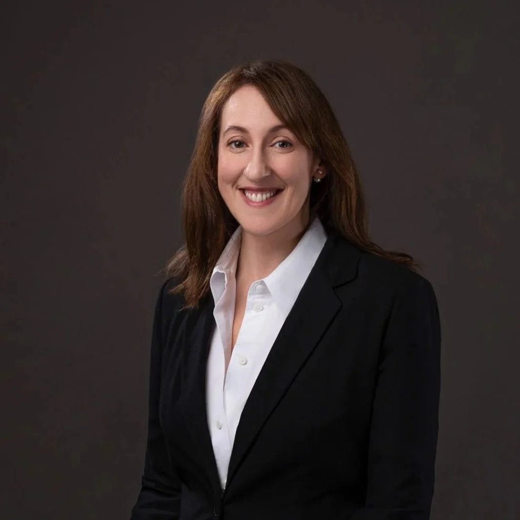 A professional woman with shoulder-length brown hair, wearing a black blazer and white collared shirt, smiling at the camera against a dark background.