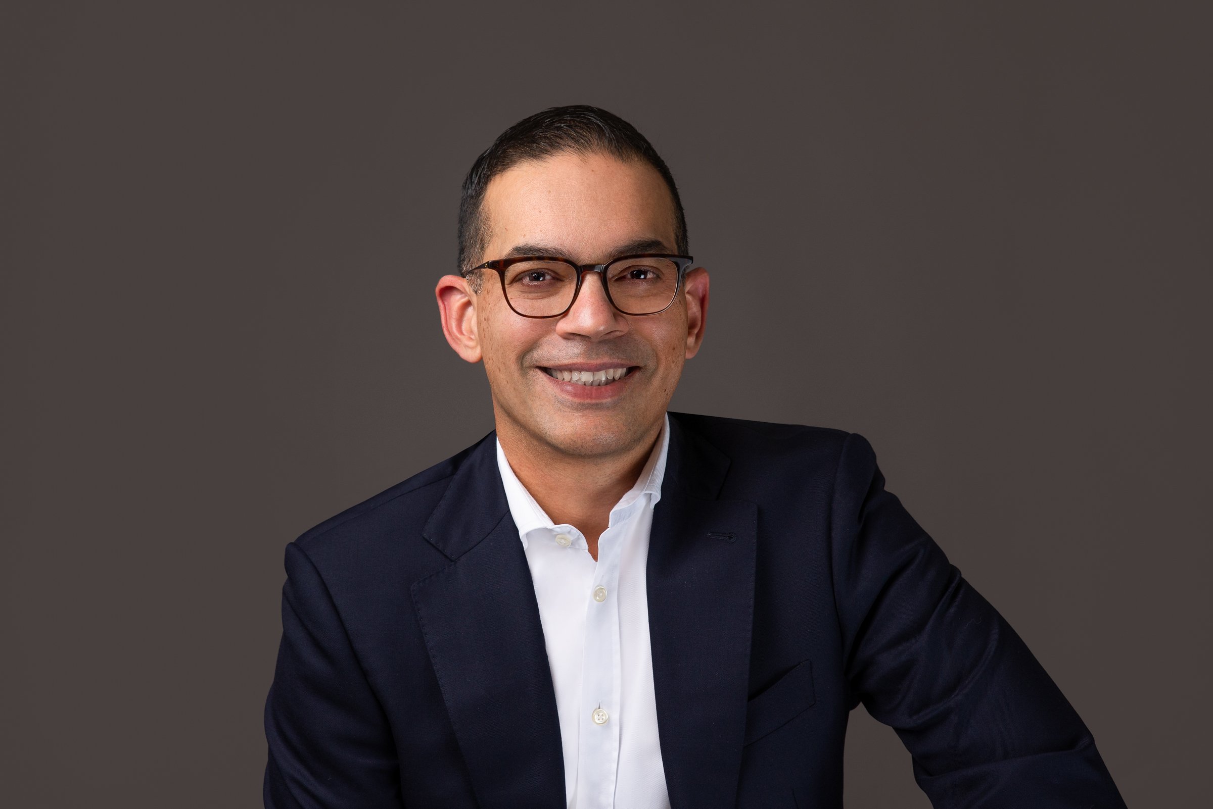Smiling man in glasses wearing a dark suit and white shirt, posed against a plain dark background.
