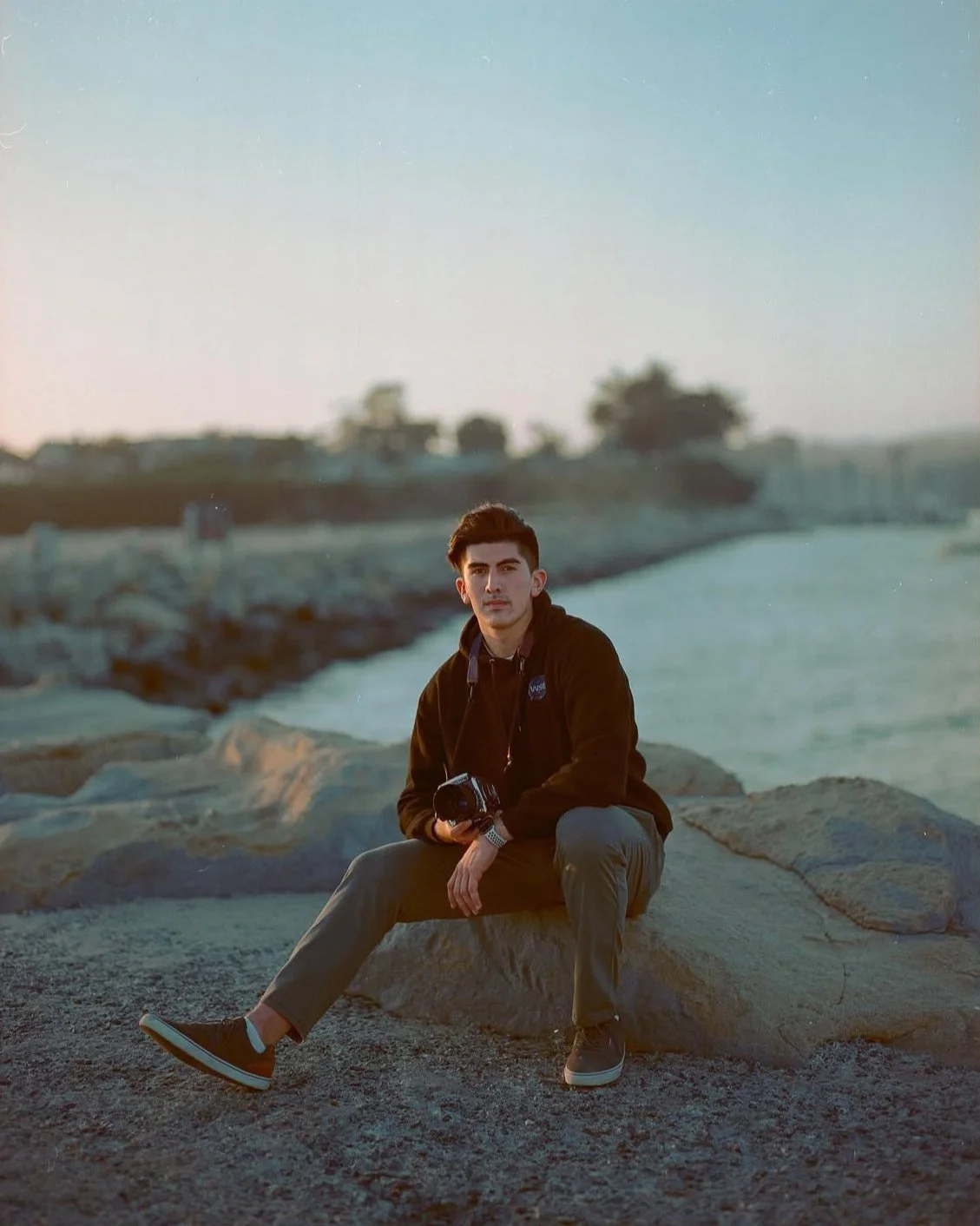 A young man sitting on a rock near the water during sunset, holding a camera, wearing a black jacket and casual gray pants.