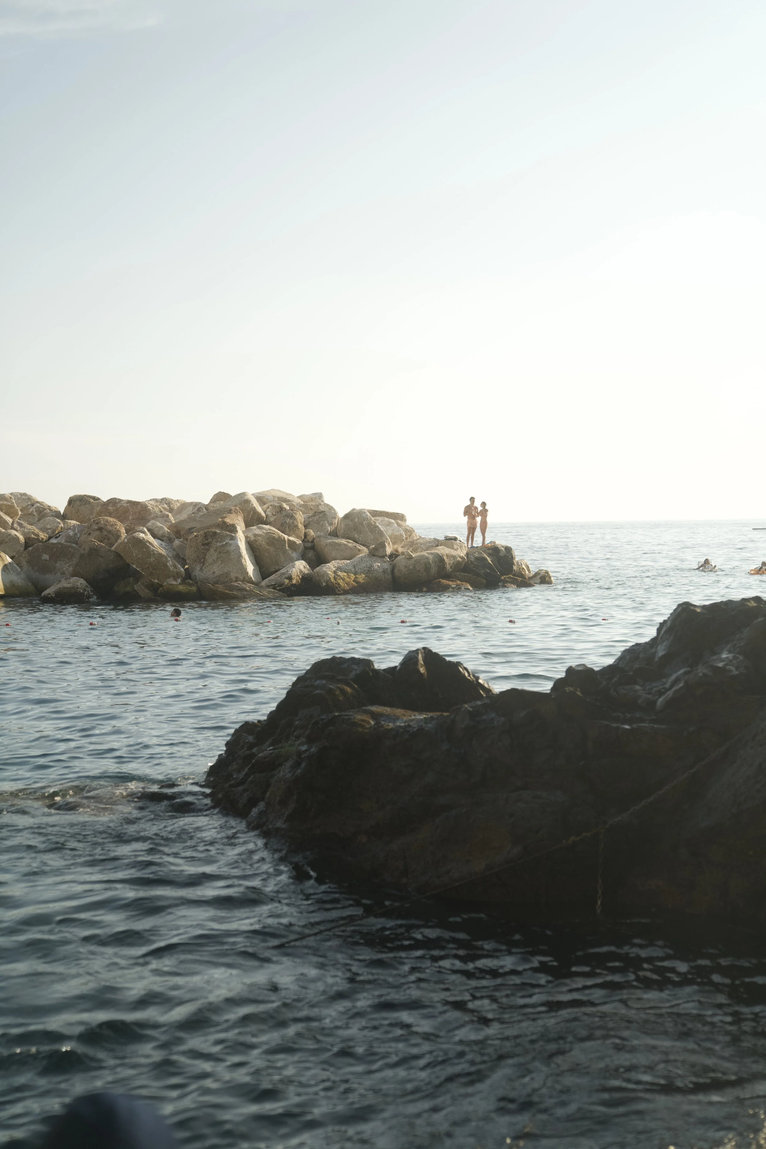 Two people standing on rocks by the ocean during sunset or sunrise, with calm water and other swimmers nearby.