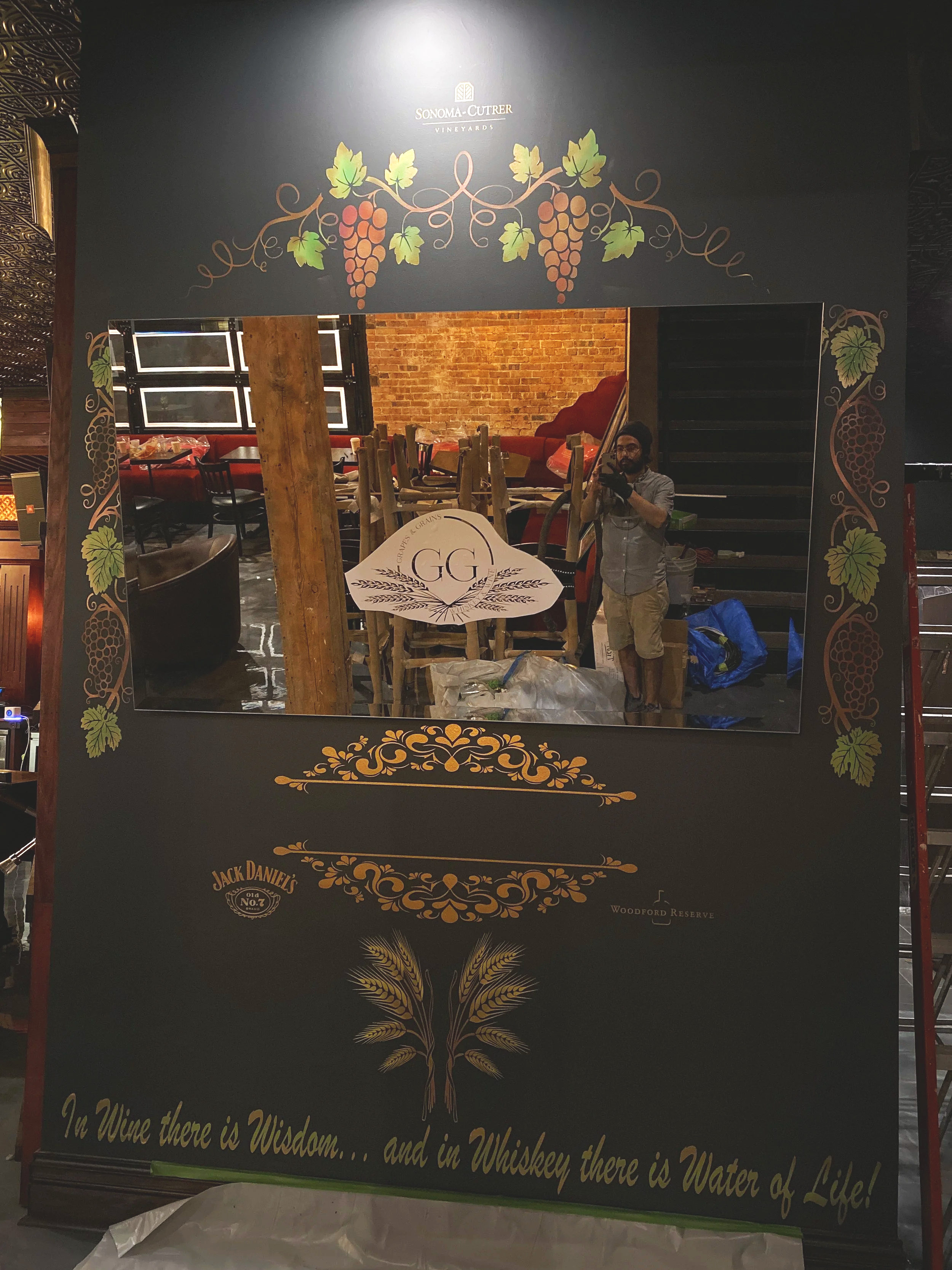 Behind-the-scenes shot of a hand-painted grapevine mural and custom vinyl logo install inside The Grape & Grain lounge.