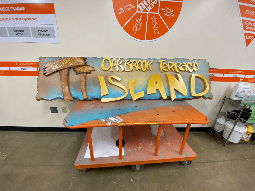 3D hand-carved foamboard retail sign with wood-textured posts and bold yellow lettering reading “Oakbrook Terrace Island,” installed in a Home Depot store.