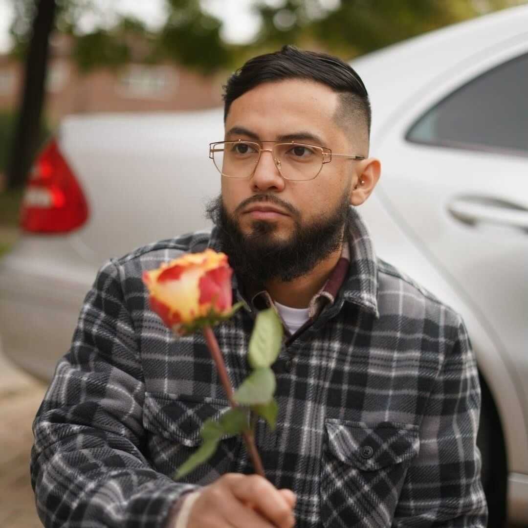 A man with glasses and a beard holding a rose in front of him outdoors.