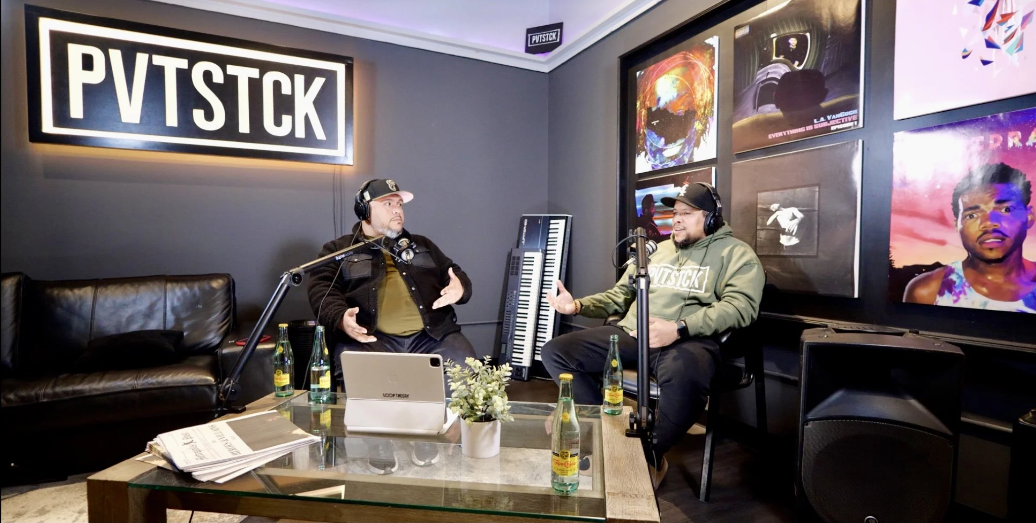 Two men wearing headphones engaged in conversation during a podcast, with one gesturing with hands and the other listening, in a decorated room with posters and artwork on the walls.