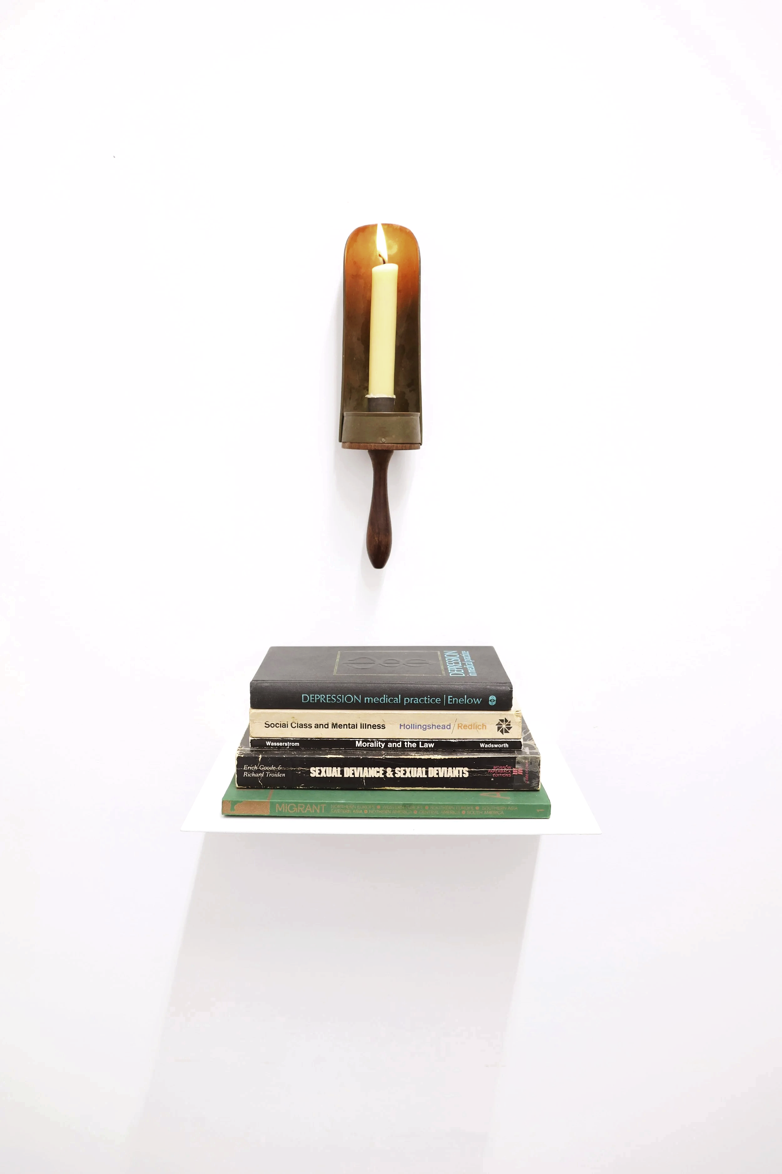 A assemblage of a stack of five books: social class, depression, morality, on a white shelve with a wall-mounted candle holder: chamber stick above. The candle holder has a lit candle and a wooden handle.