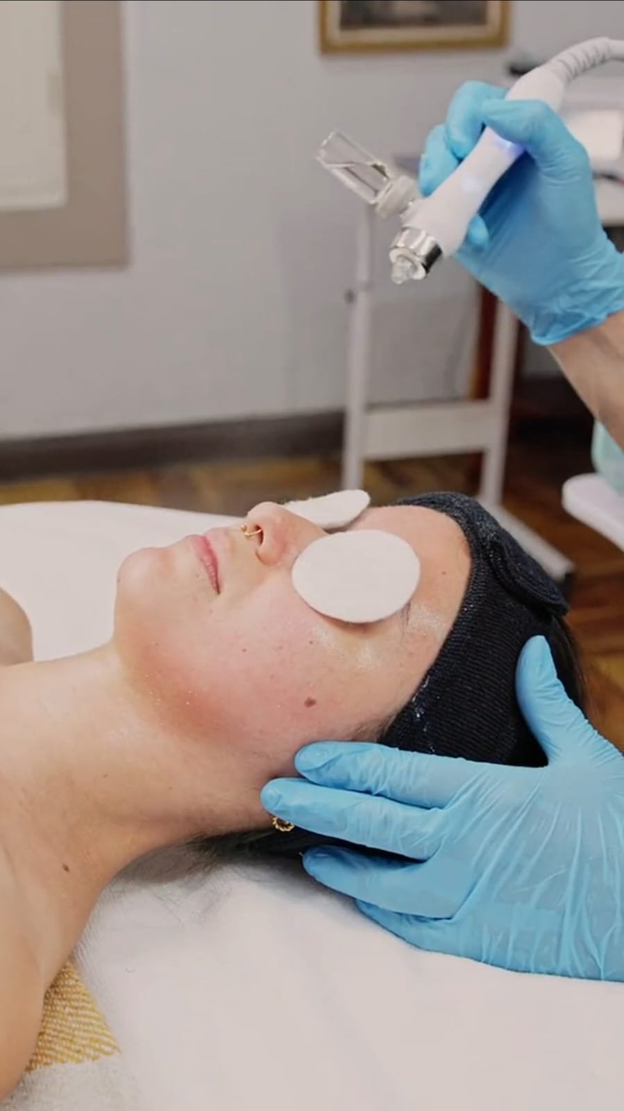 A woman receiving a facial treatment with cotton pads on her face, while a skincare professional holds a device above her face in a clinical setting.