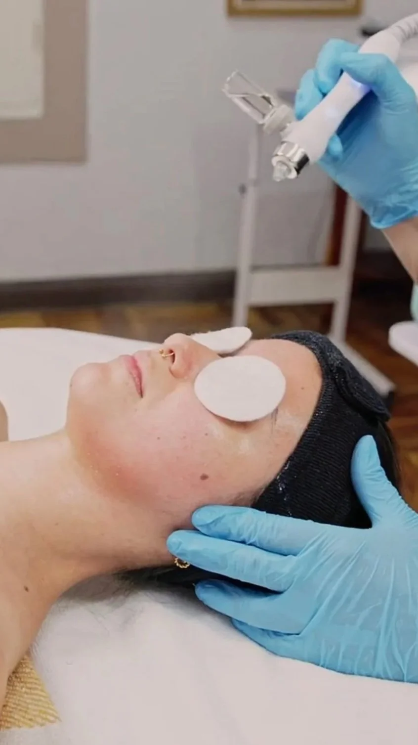 A person receiving a facial treatment with eye pads, wearing a black headband, lying on a treatment bed, and a skincare professional in blue gloves holding a syringe.