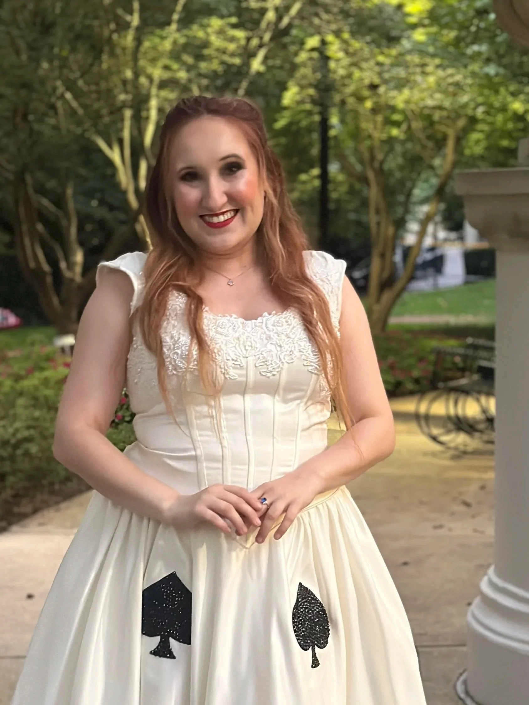 A woman with long red hair wearing a white dress with lace details and black spade symbols on the skirt, smiling outdoors in a park with trees and greenery in the background.