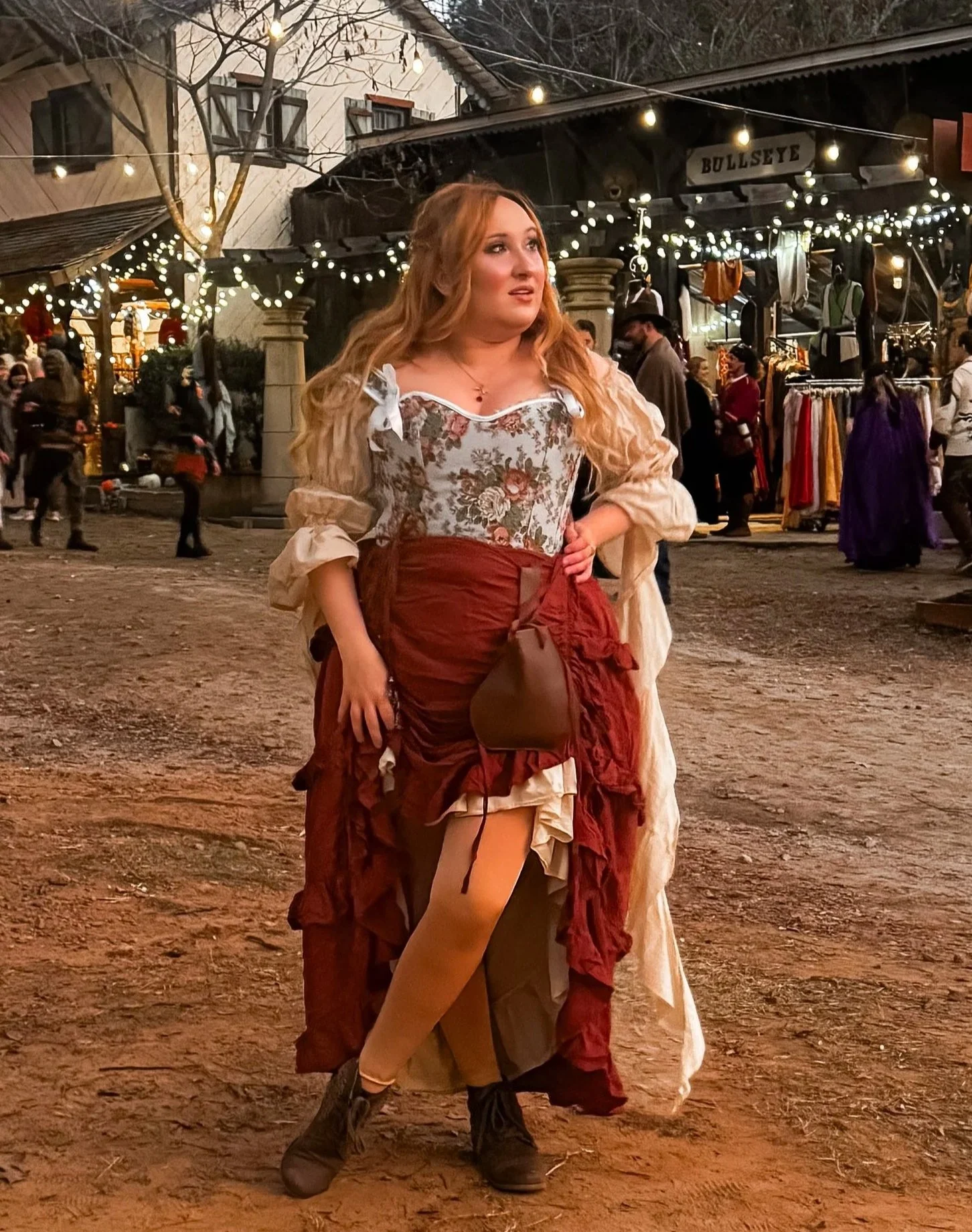 A woman in vintage-style clothing standing at a lively outdoor market or fair with string lights and temporary shops.