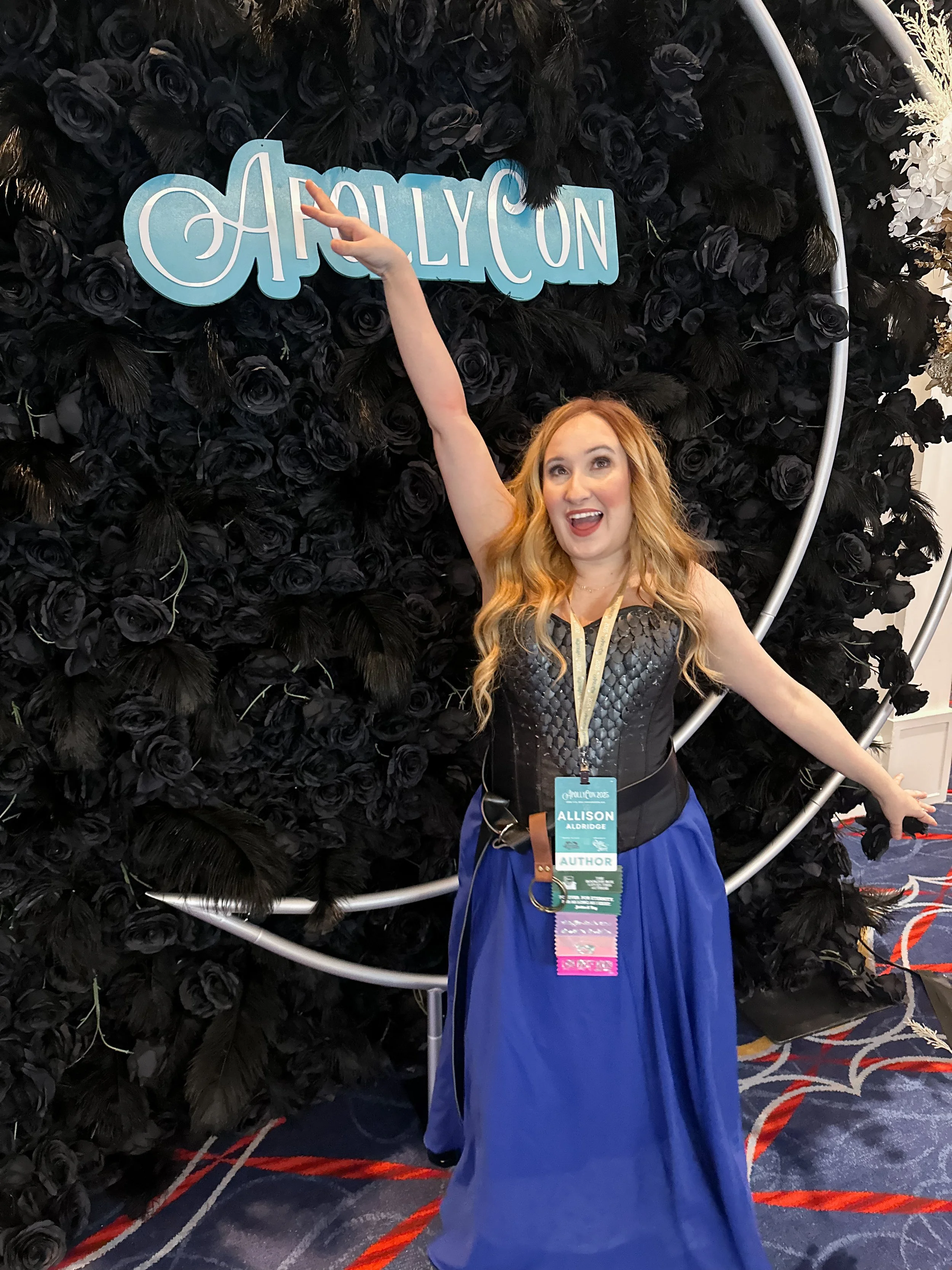 A woman with long, wavy red hair, wearing a strapless black top and a long blue skirt, stands in front of a black floral wall with a large circular metal frame. She is pointing upwards with her right hand towards a sign that reads 'Apollyon' and smiling, with multiple event badges around her neck.
