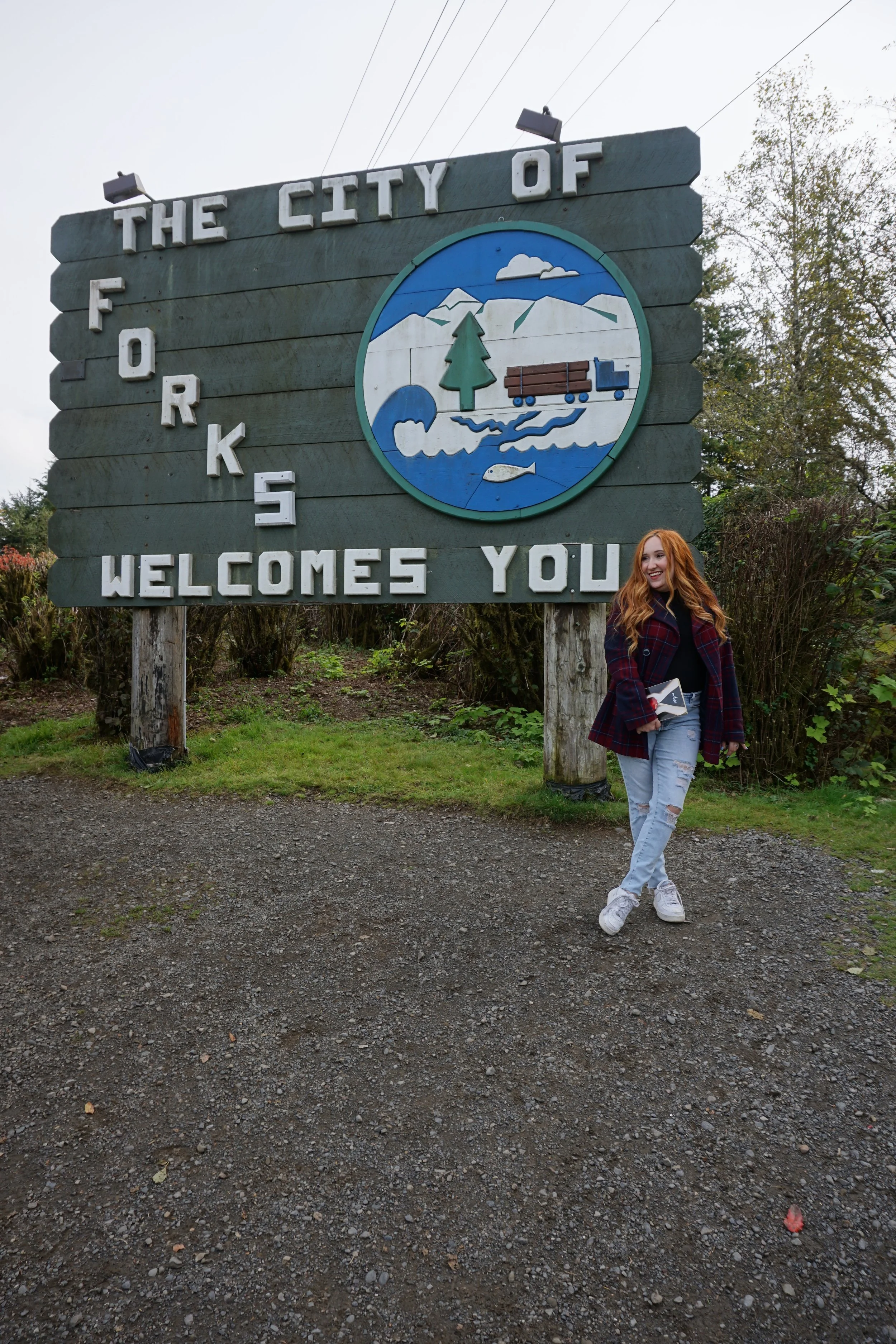 A woman with red hair in casual clothing standing in front of a large wooden welcome sign for the city of Forks, featuring a logo with mountains, trees, a river, and a fish.