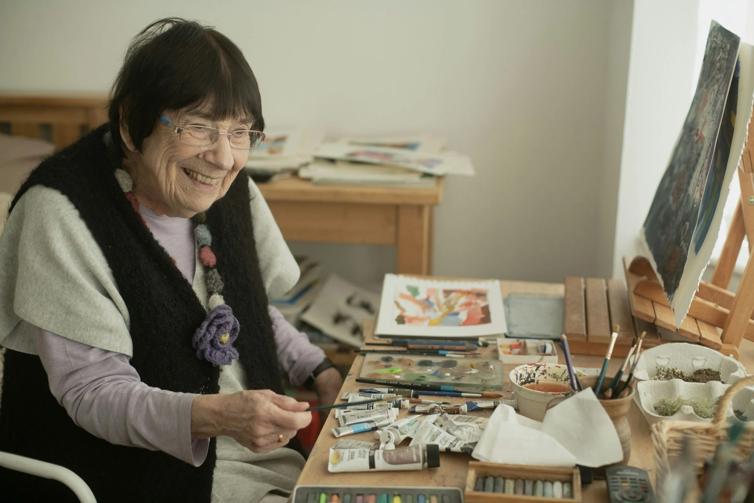An elderly woman with glasses and a black knitted vest smiling while painting at her art setup in a well-lit room.