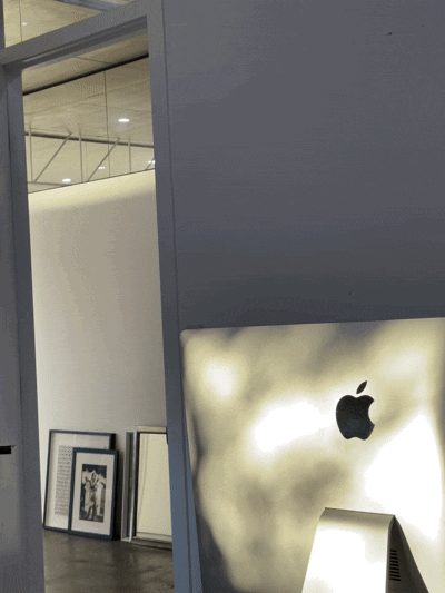 Apple logo on a backlit display inside an Apple store with framed pictures leaning against the wall in the background.