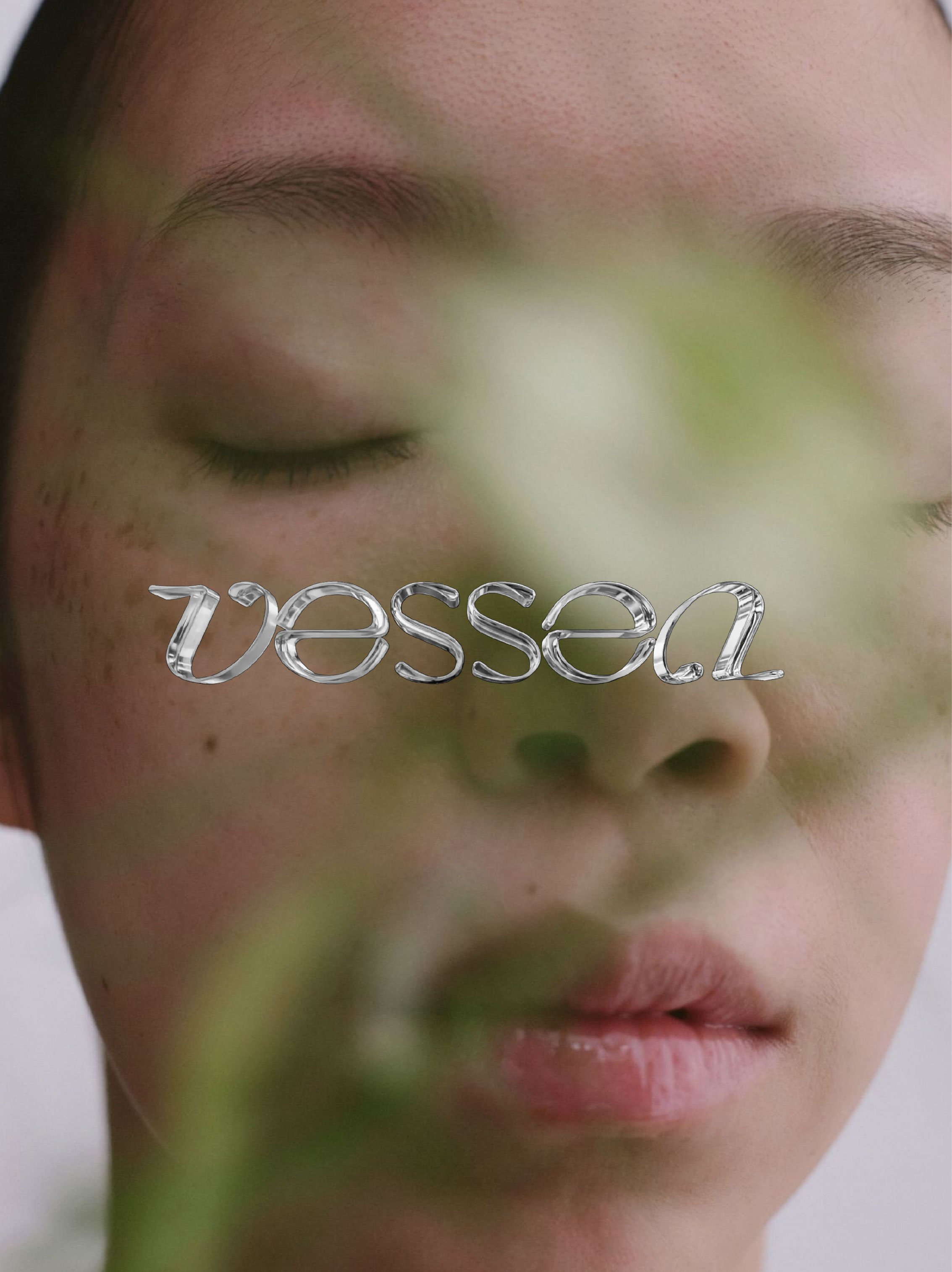 Close-up of a woman's face with a blurred green plant in the foreground and the word 'vessa' overlayed in metallic font across her nose.