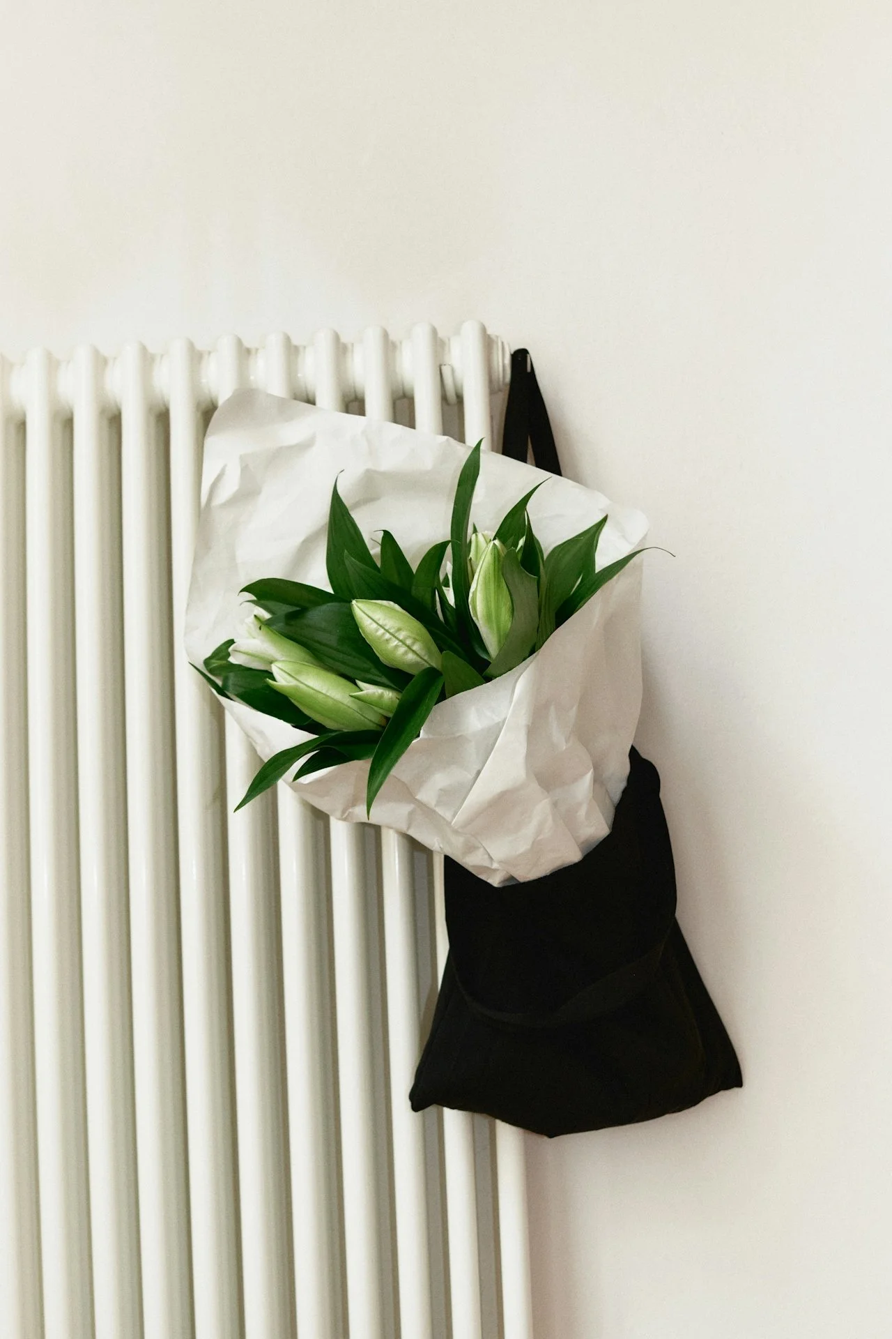 Bouquet of white lilies with green leaves wrapped in white paper hangs on a white radiator against a plain white wall.