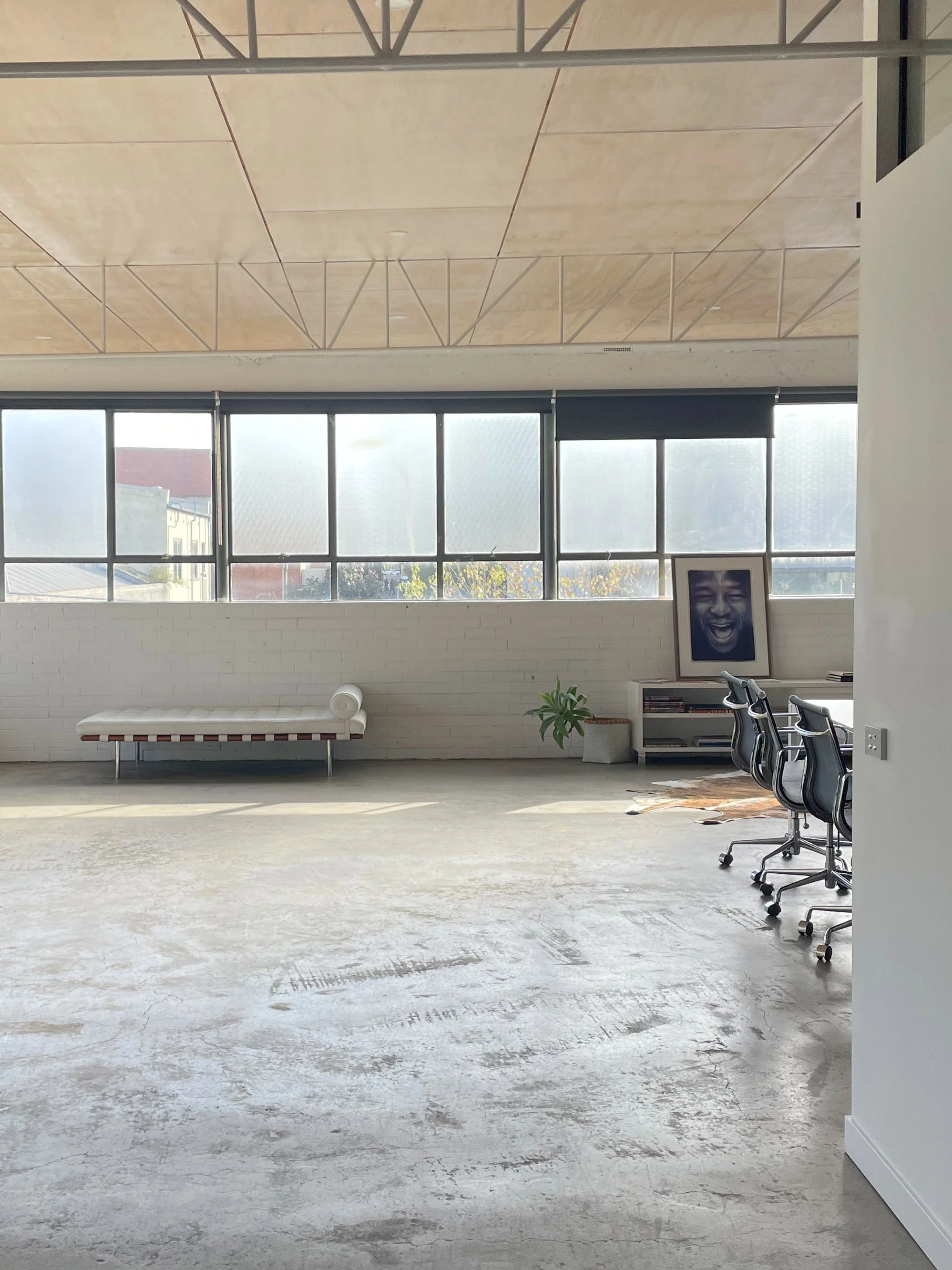 Modern office or studio space with large windows, white brick wall, minimal furniture including a bench, plant, framed photo of a laughing person, black office chairs, and a rug. The ceiling has a unique geometric design.