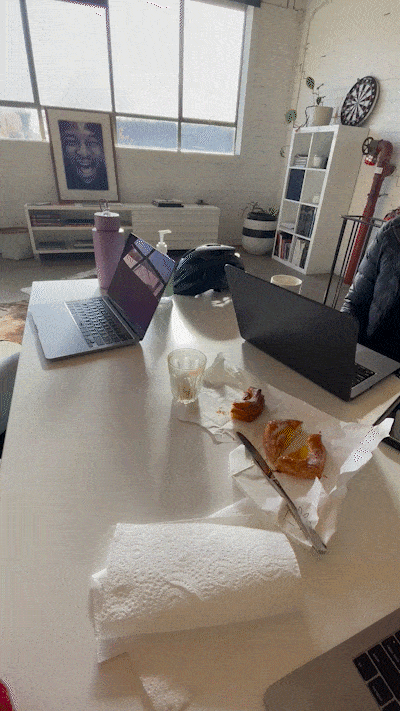 A white table with two laptops, a purple water bottle, a glass with a drink, some food, a piece of paper towel, and a plastic bag with food. In the background, a window, a framed photo, shelves with books and decor, and a dartboard on the wall.