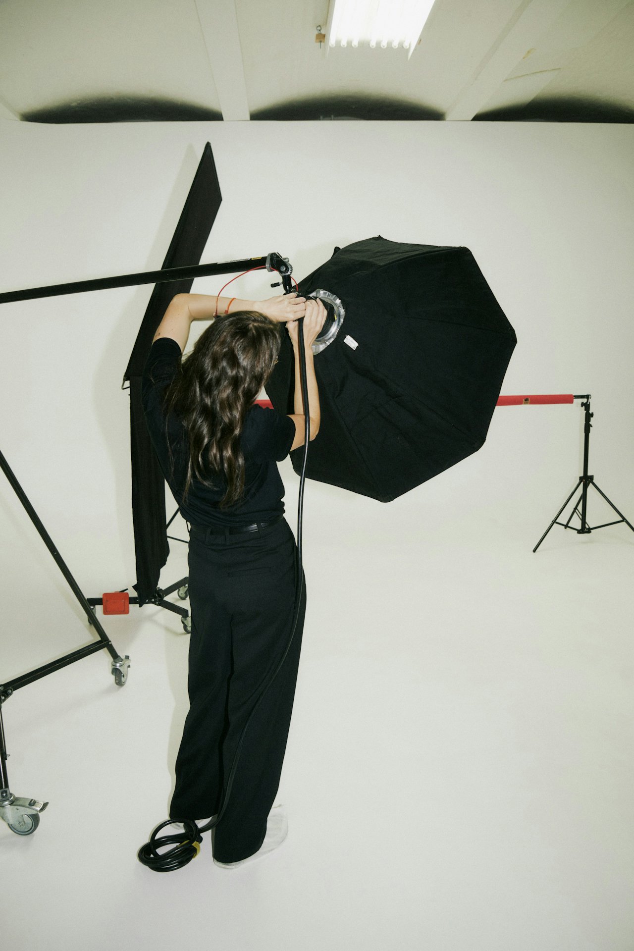 A woman preparing photography equipment in a studio with a large umbrella light and a plain white backdrop.