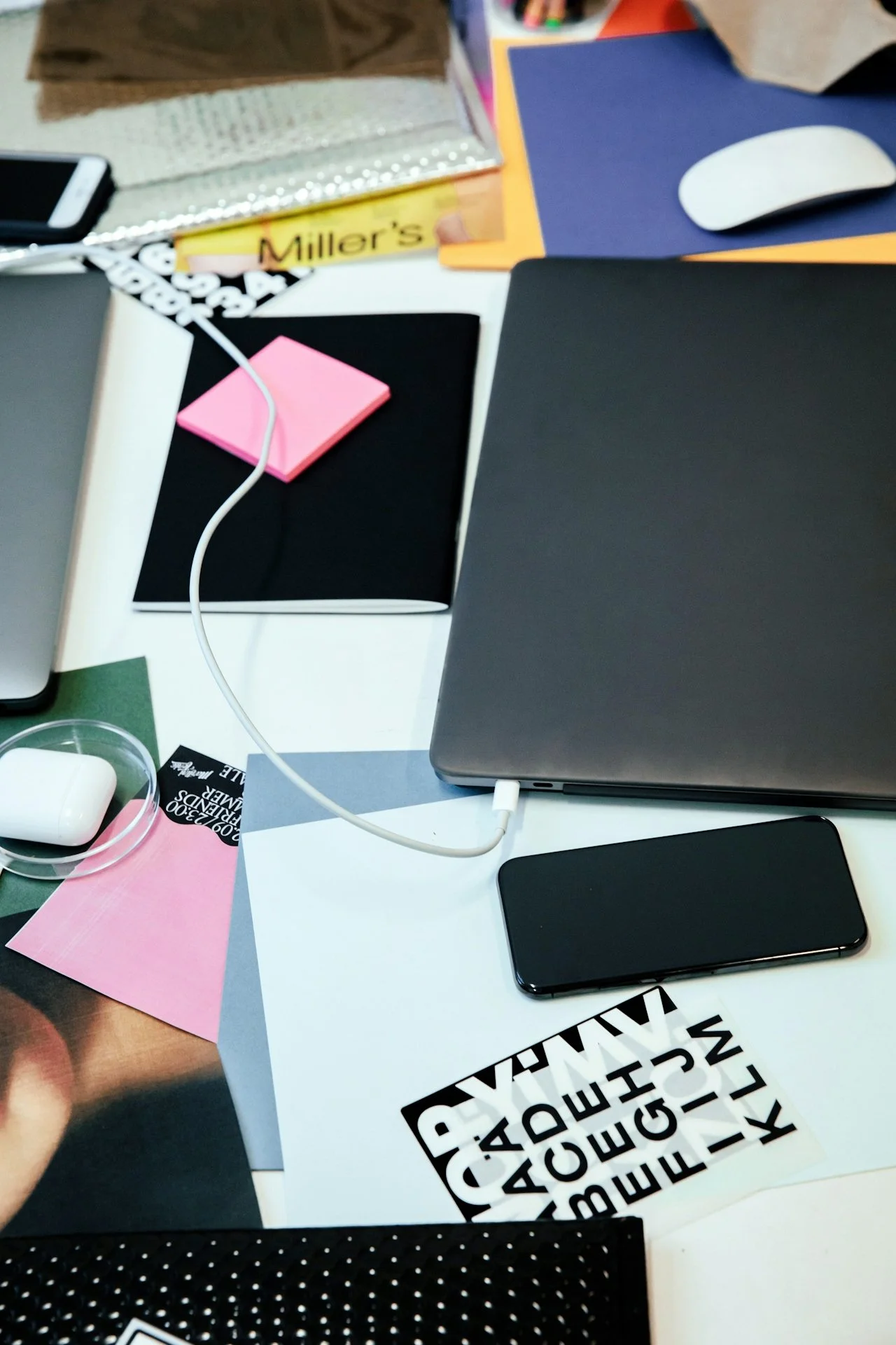 Cluttered workspace with a laptop, smartphone, wireless earbuds, sticky notes, paper, and various office supplies on the desk.