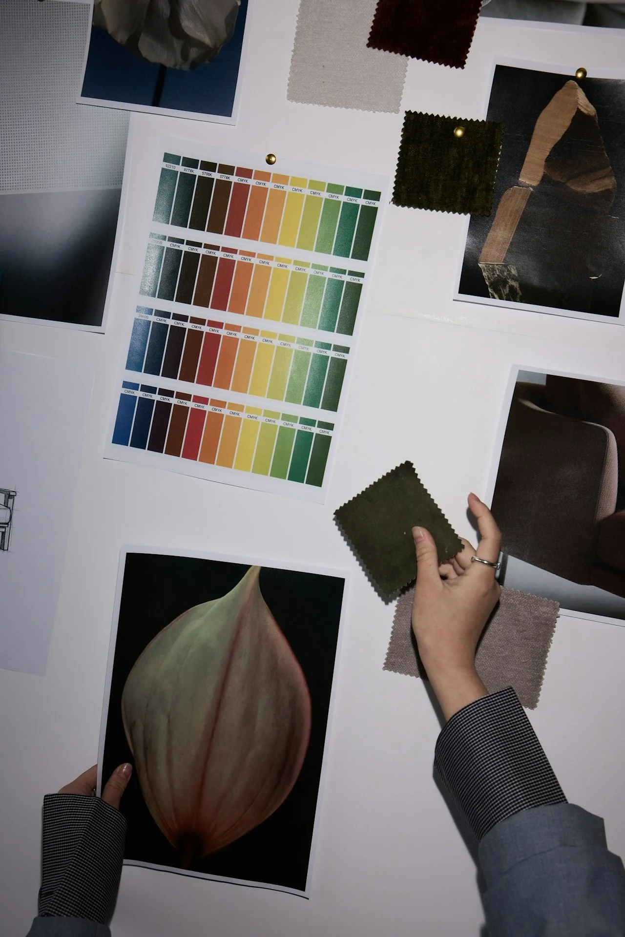 A person holding fabric sample swatches over a workspace with color charts, fabric swatches, and printed images of a plant and a dark reflective surface.
