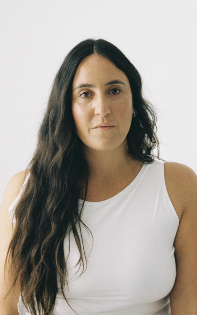 A woman with long, wavy dark hair, wearing a white sleeveless top, standing against a plain light-colored background.