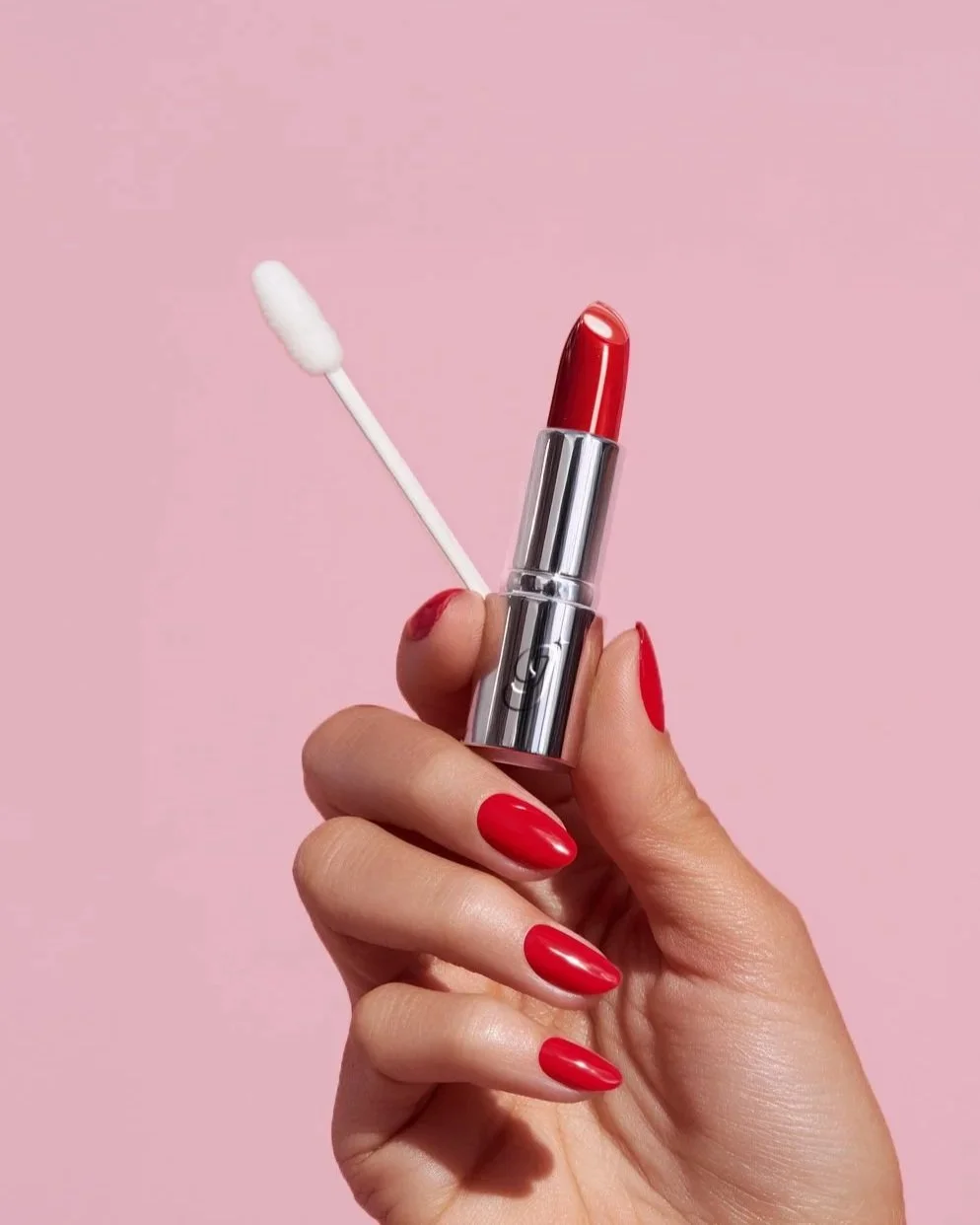 A hand with red nail polish holding a lipstick with a red cap and a white applicator against a pink background.