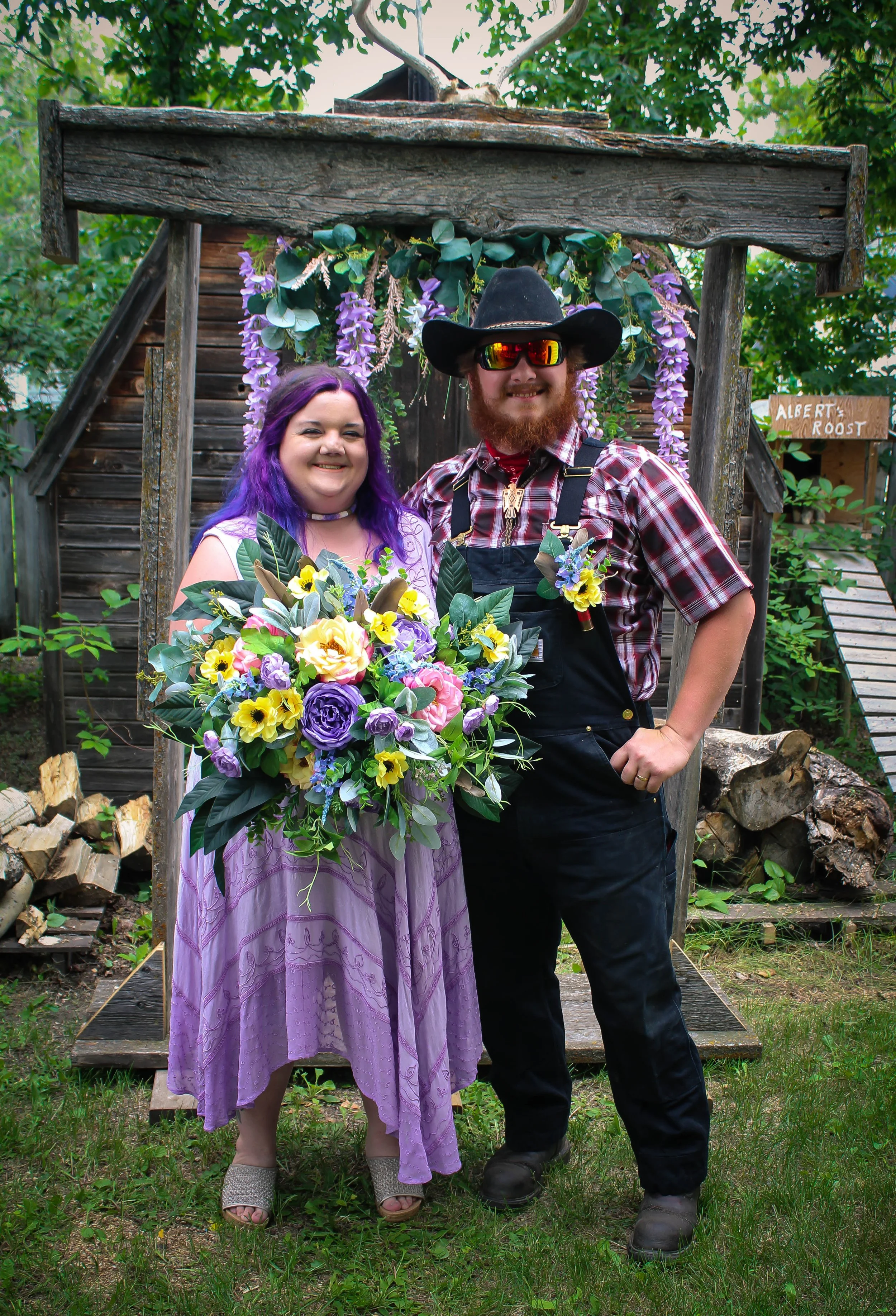 A woman with purple hair and a man with a beard wearing a black cowboy hat, sunglasses, plaid shirt, and overalls. They are holding a large bouquet of colorful flowers. They are standing outdoors in front of a rustic wooden structure decorated with purple wisteria flowers, surrounded by trees and logs.