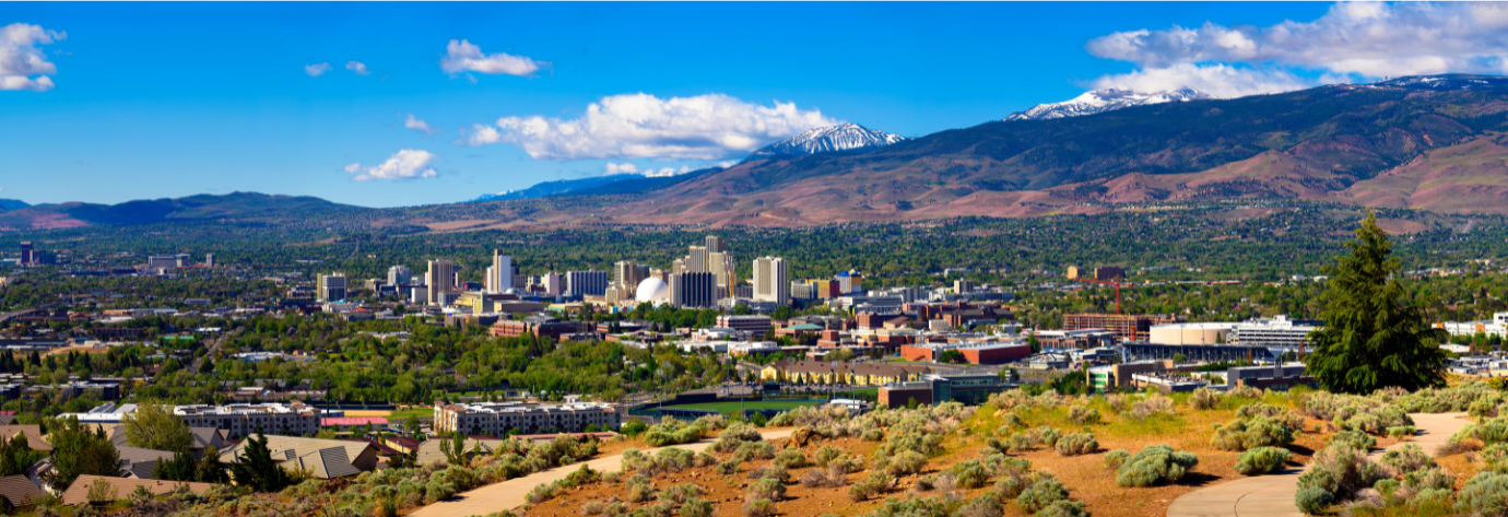 Panoramic view of a Reno and Sparks, NV with green trees, and mountains with snow in the background under a partly cloudy sky.