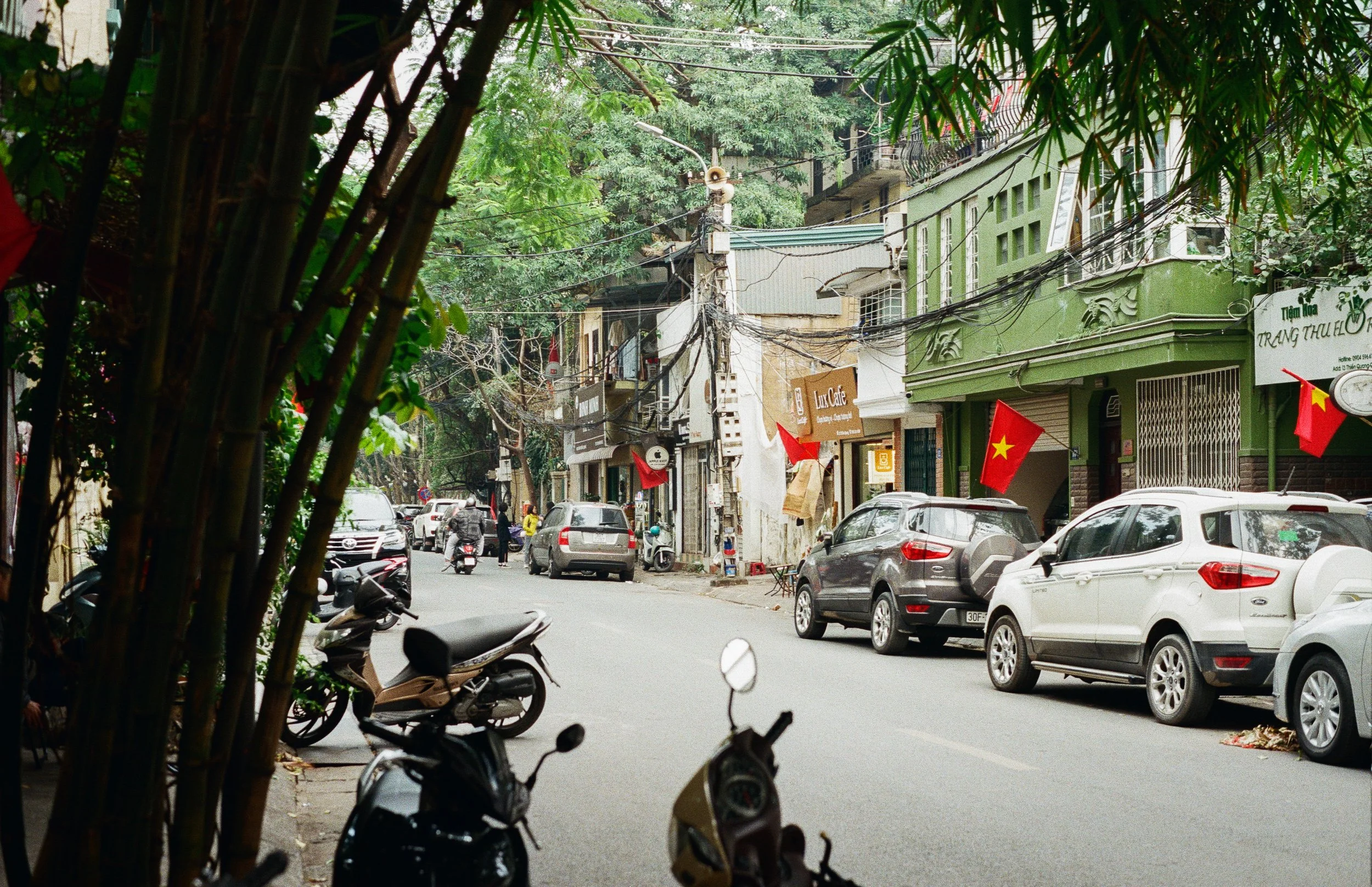 Street scene in Vietnam with parked cars, motorbikes, buildings with storefronts, and Vietnamese flags hanging outside.