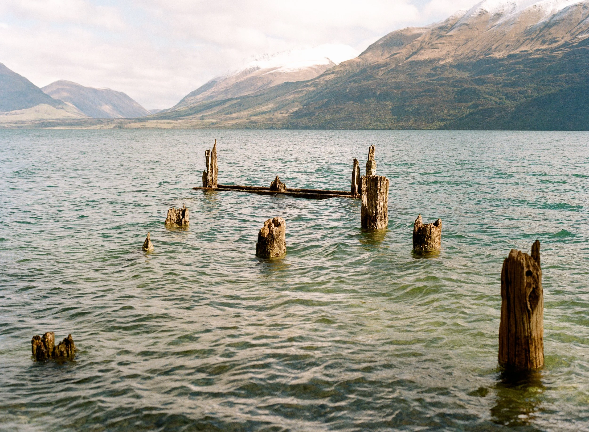 Old wooden pilings and remains of a dock protrude from a calm body of water, with mountains in the distance and a cloudy sky overhead.