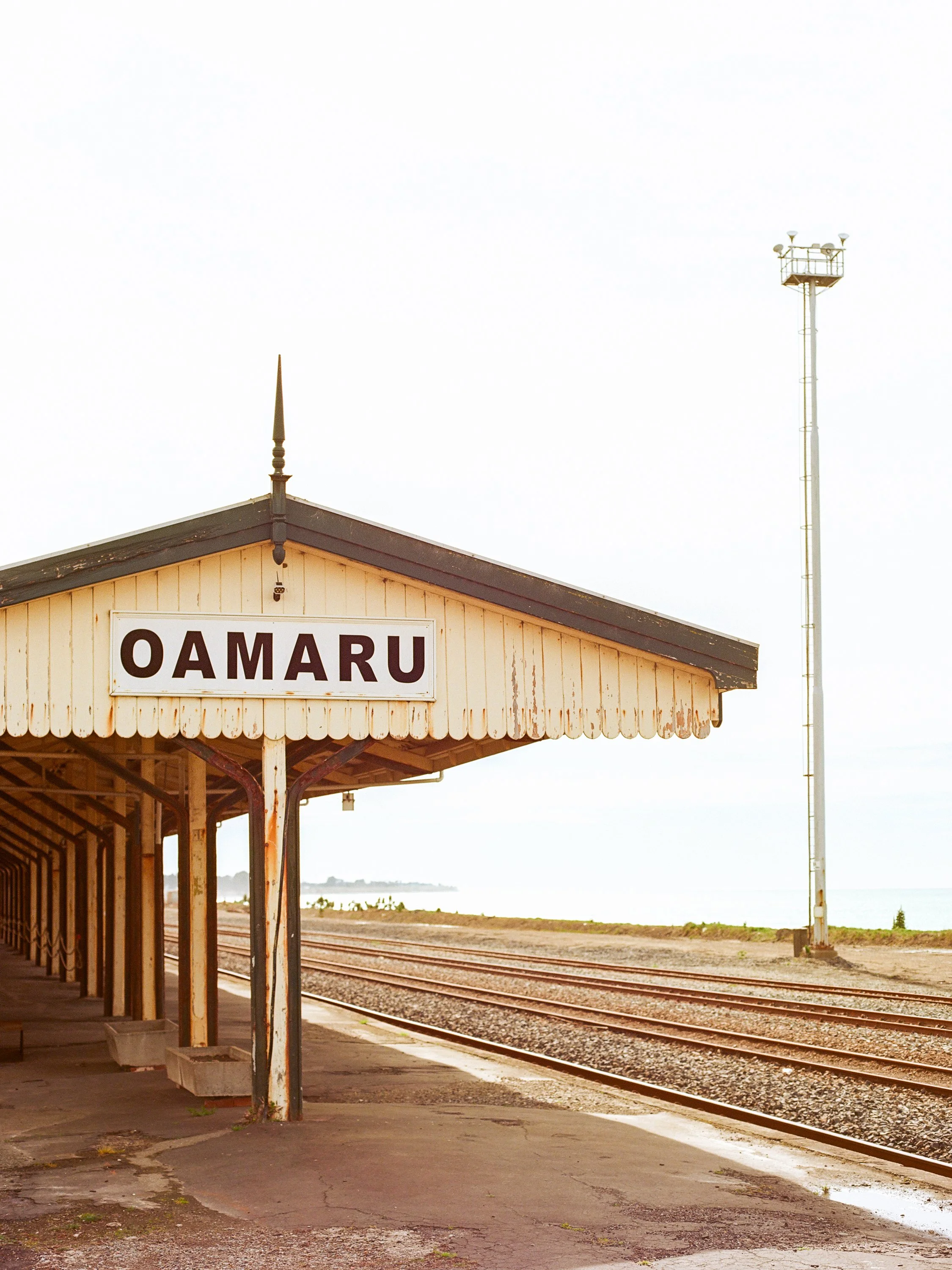 A vintage train station platform with a sign that reads 'Oamaru', overlooking train tracks and the ocean in the background.
