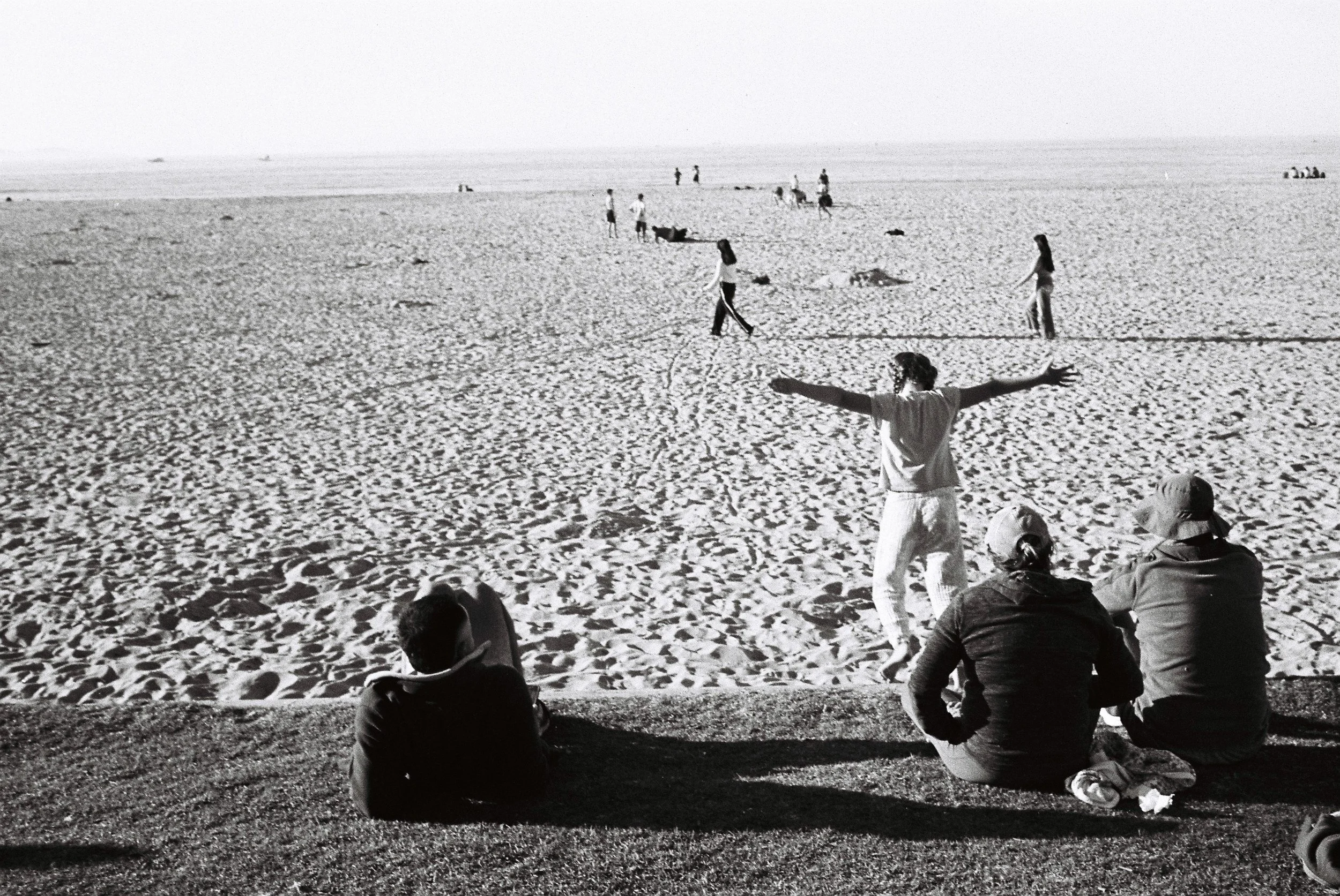 People on a sandy beach, with some sitting and others walking or playing, and the ocean in the background.