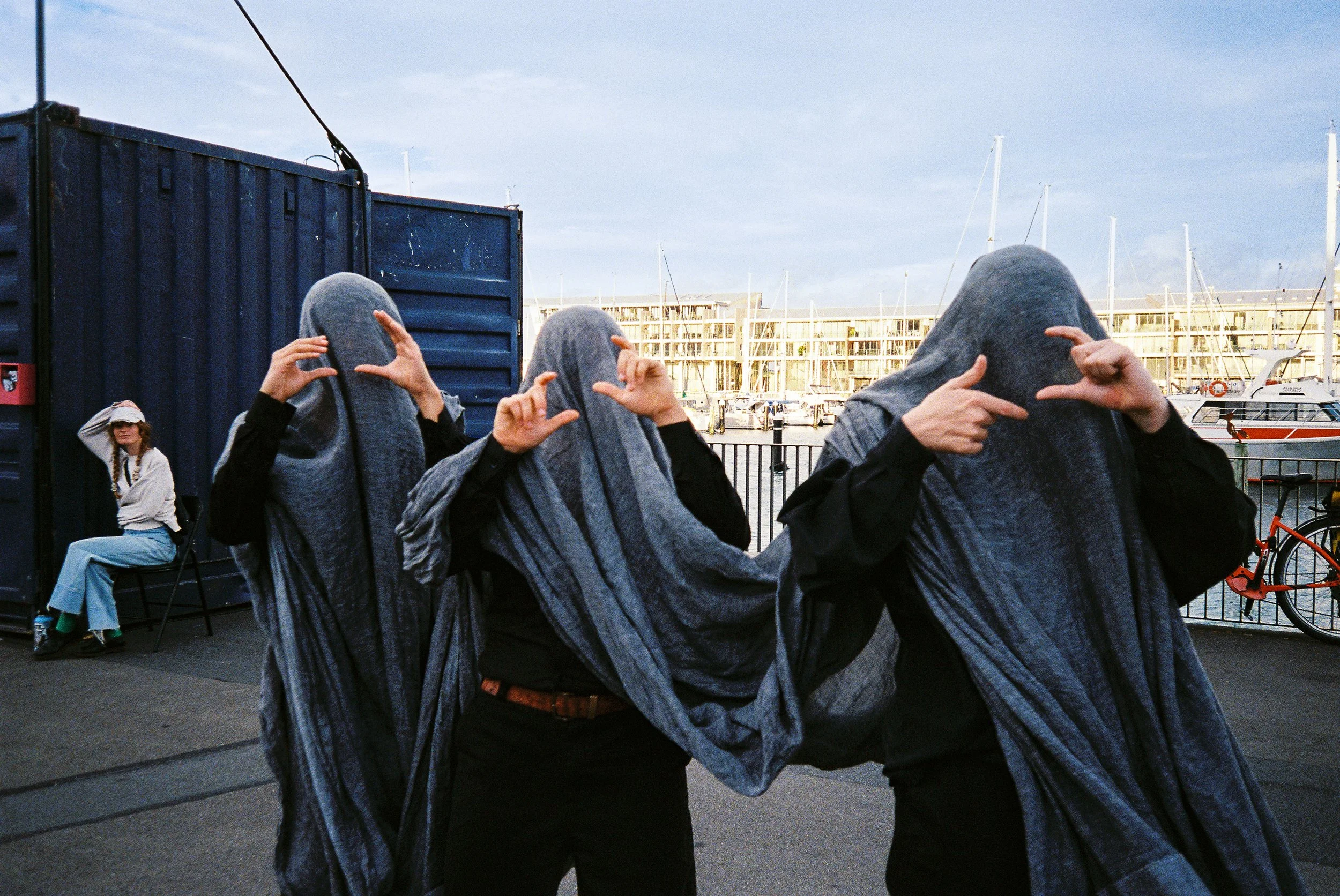 Three people with their heads covered by gray striped fabric, standing near a marina with boats and buildings in the background. A woman sitting on a chair is seen to the left, wearing sunglasses and a white hoodie.