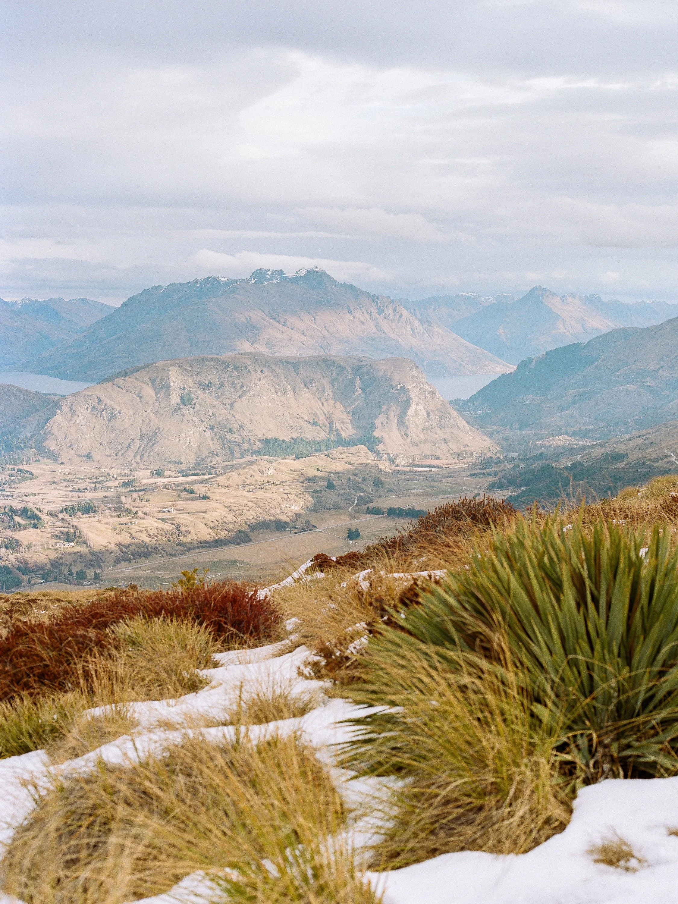 Mountain landscape with snow patches in the foreground, tall grasses, rolling hills, and distant mountains under a cloudy sky.