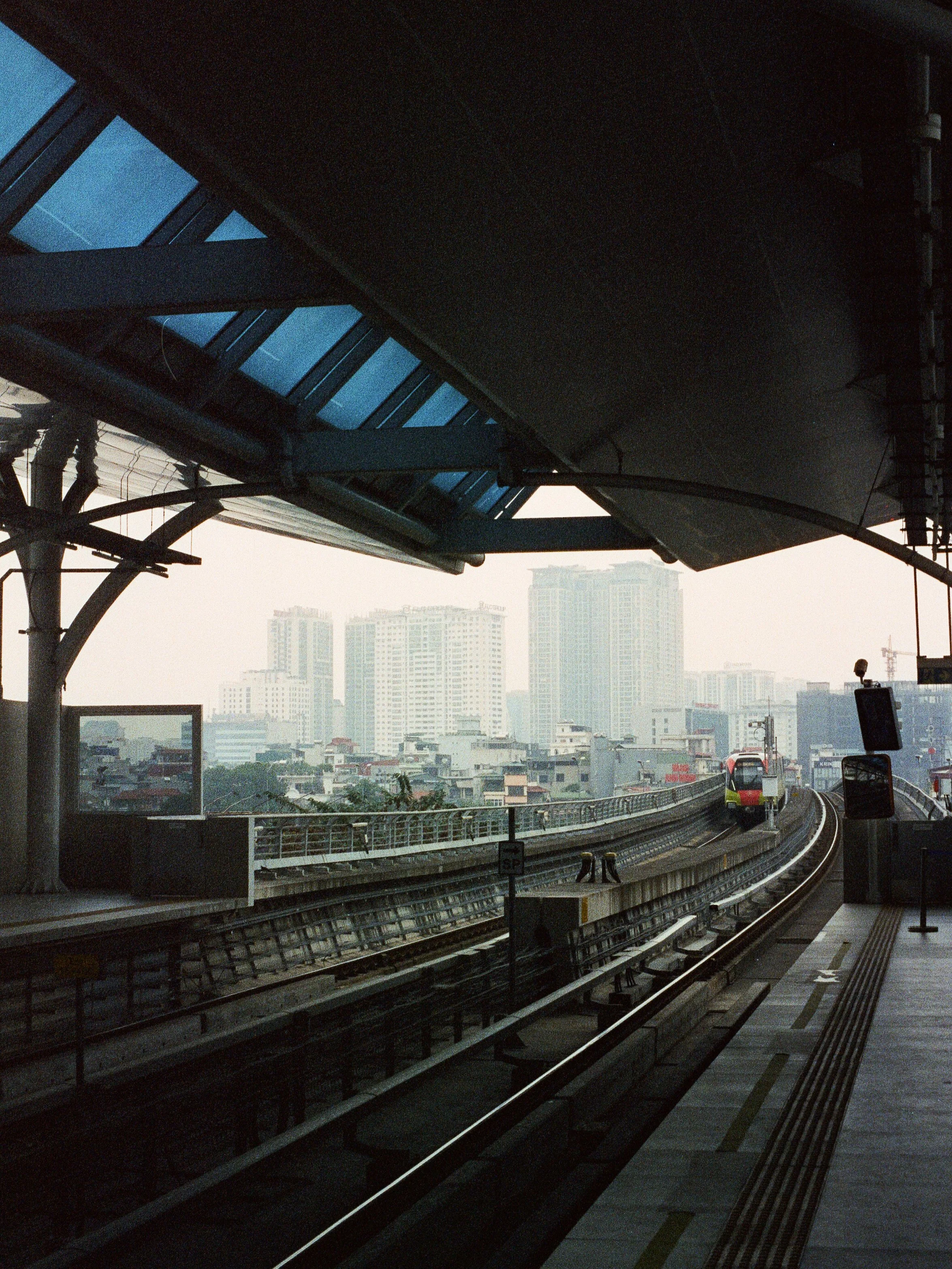 An elevated train station with a train approaching on the tracks, high-rise buildings in the background, and a platform with safety markings.