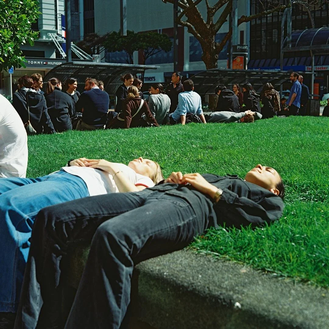 People laying on the grass with their eyes closed in an urban park, while others sit and stand in the background near storefronts and trees.