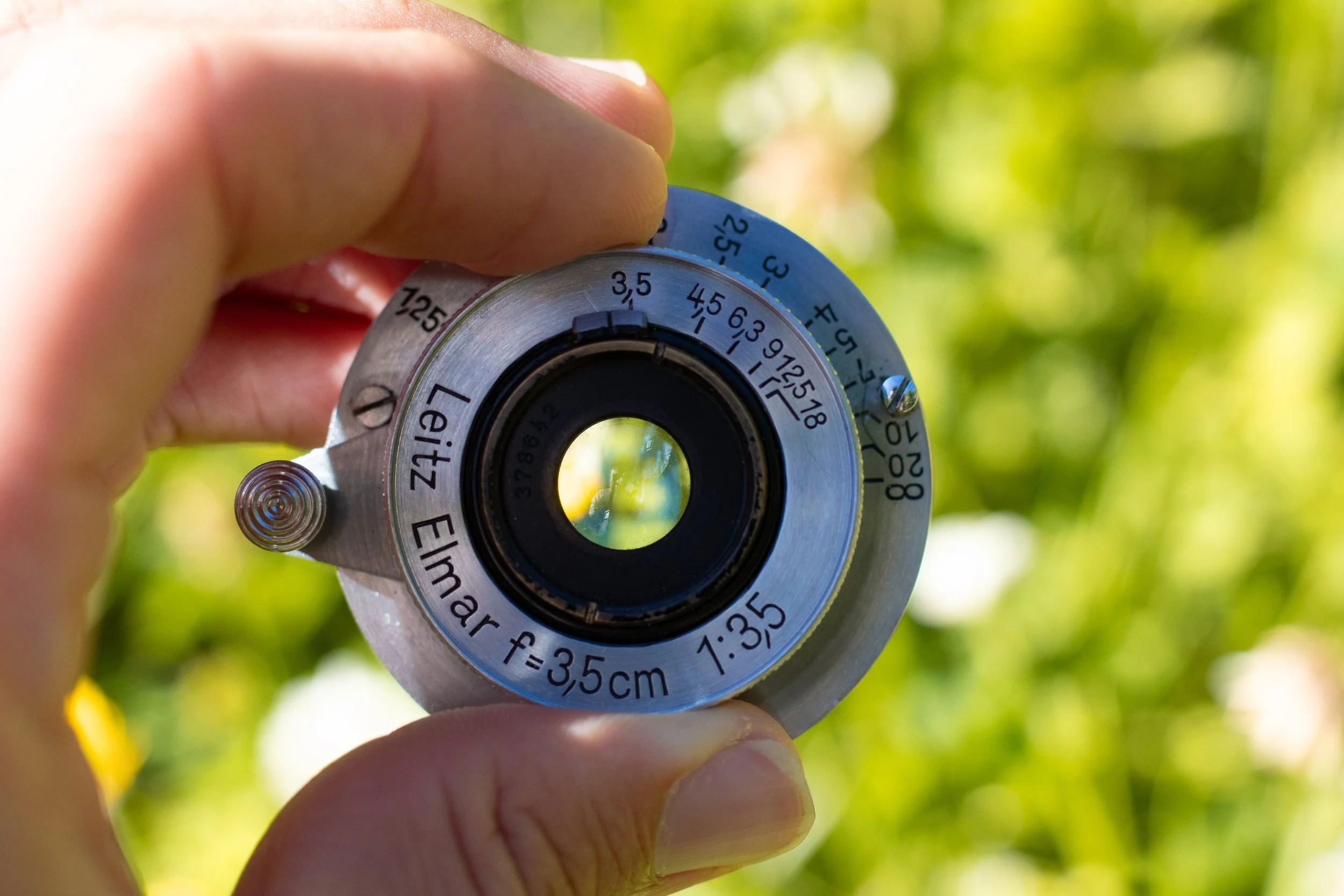Close-up of a person's hand holding a camera lens focusing device with measurement markings, against a blurred green outdoor background.