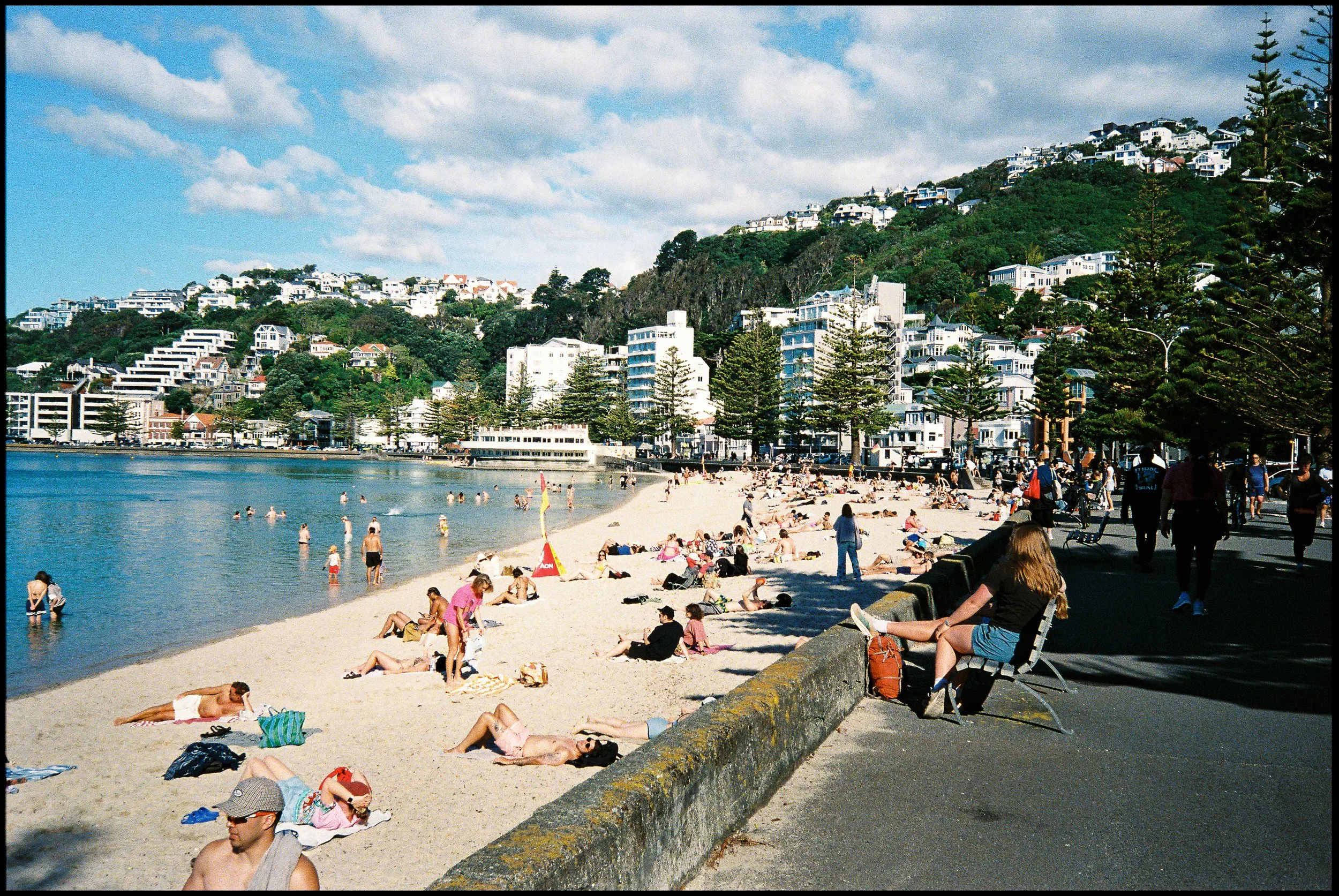 Beach scene with numerous people sunbathing and swimming, lined with waterfront buildings and green hills in the background, under a partly cloudy sky.