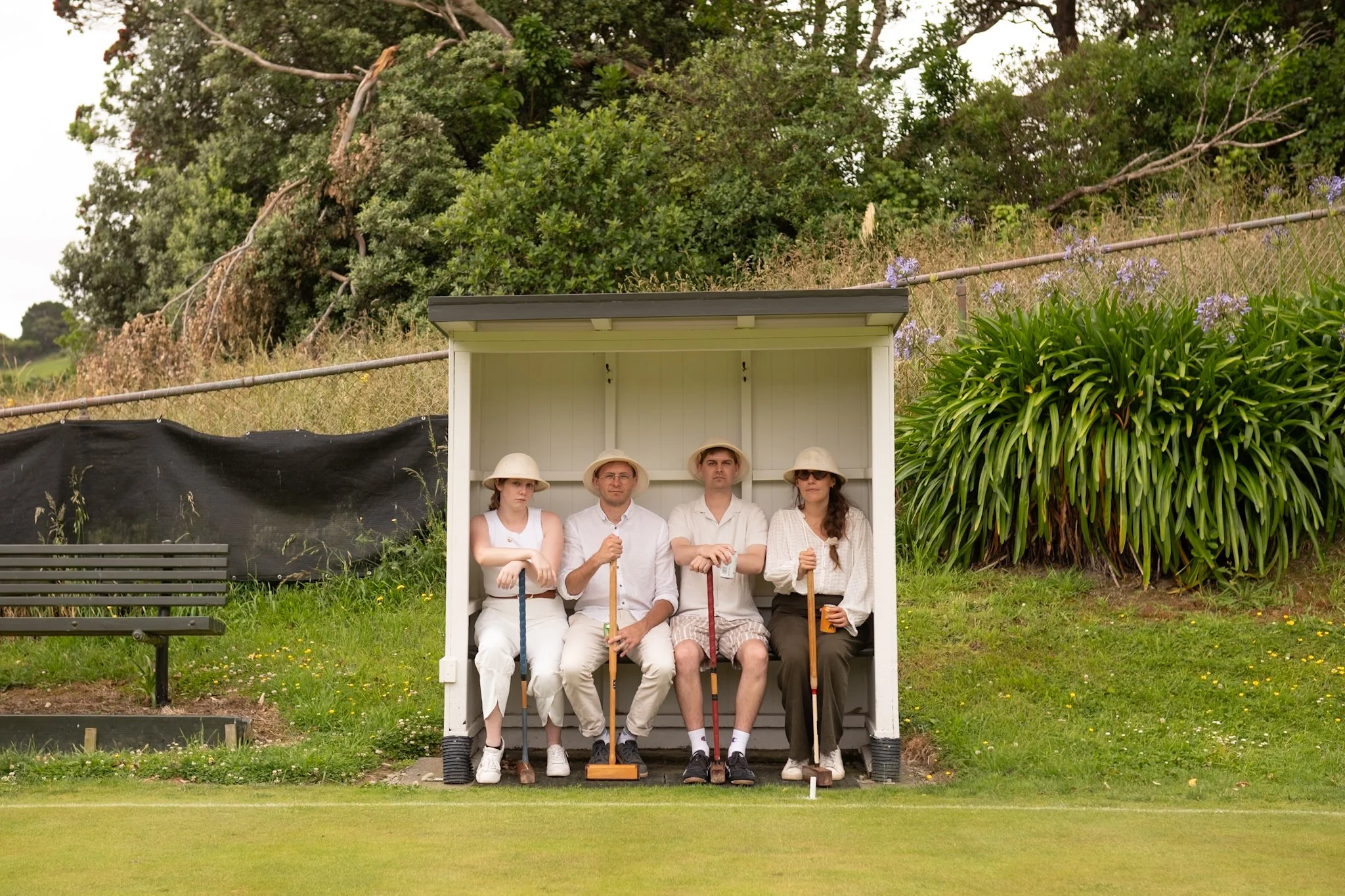 Four people sitting on a bench under a small shelter, holding croquet mallets, on a grassy field with greenery and trees in the background.