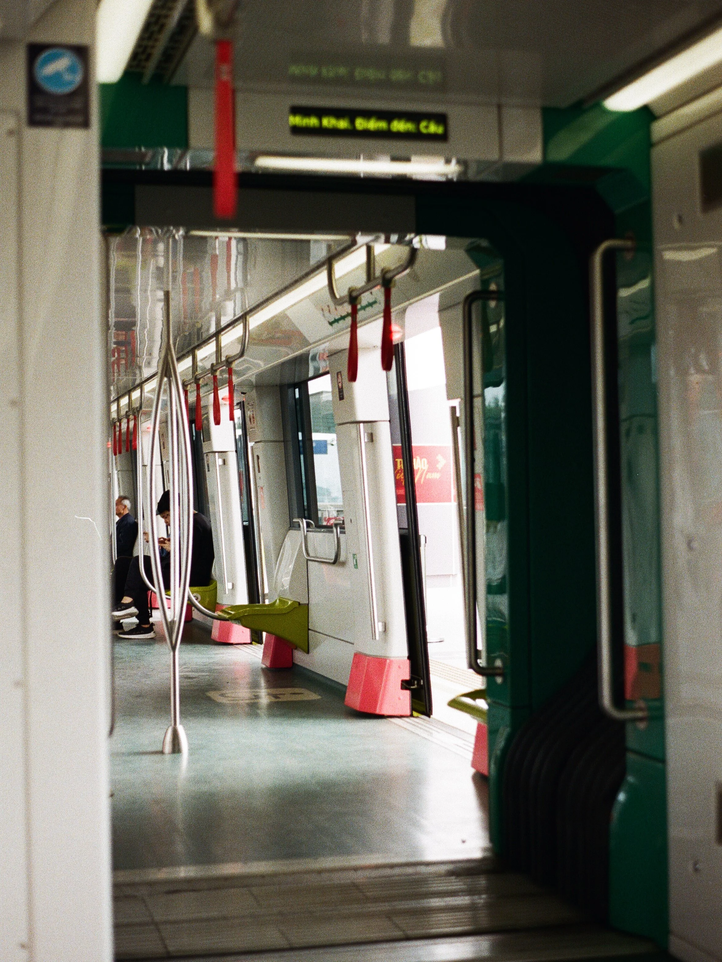 Interior of a public transit train with seats, handrails, and hanging straps, some passengers seated, with an open door showing platform outside.