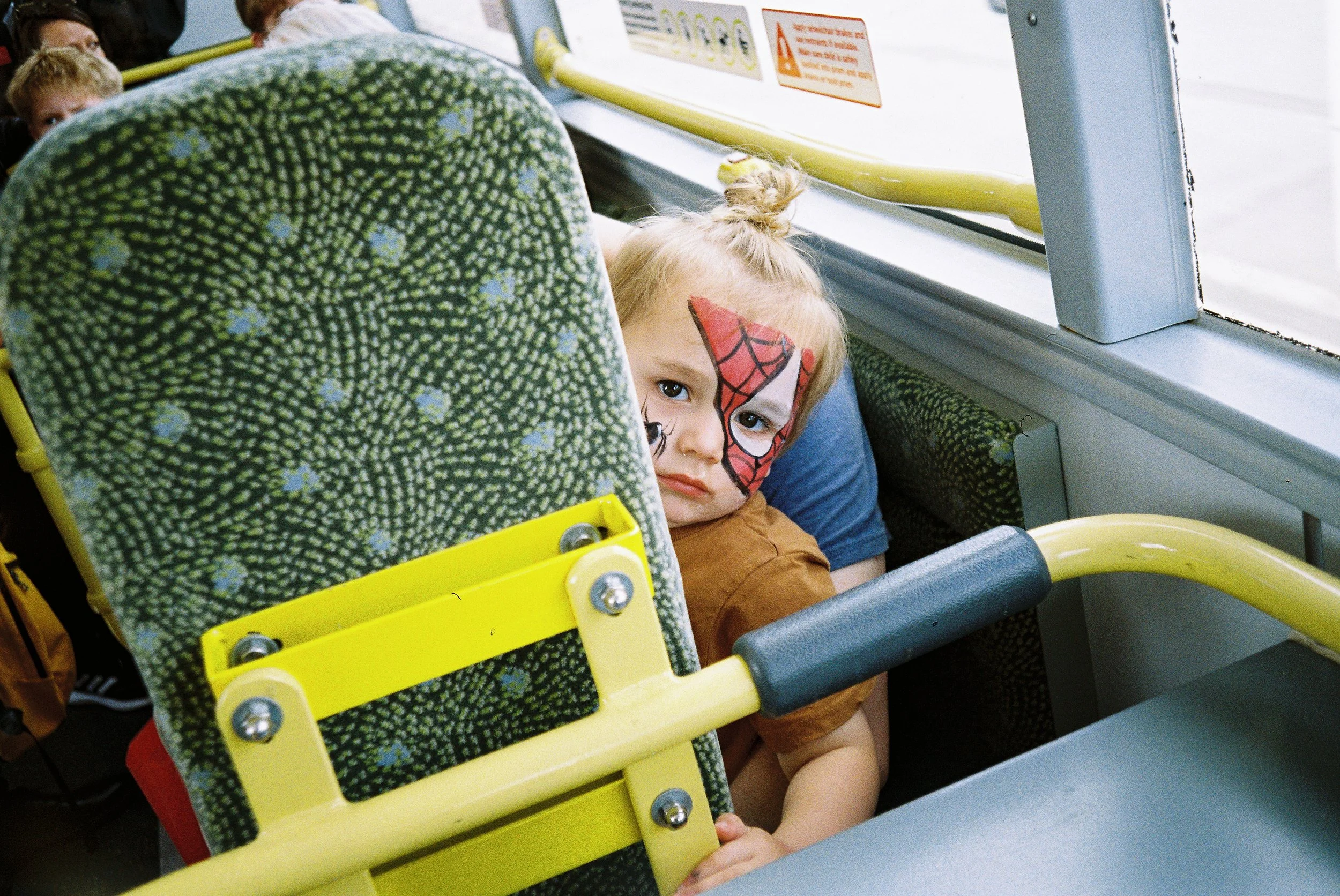 A young girl with face paint resembling Spider-Man, sitting in a yellow and green patterned bus seat, resting her head on the side window with other passengers in the background.