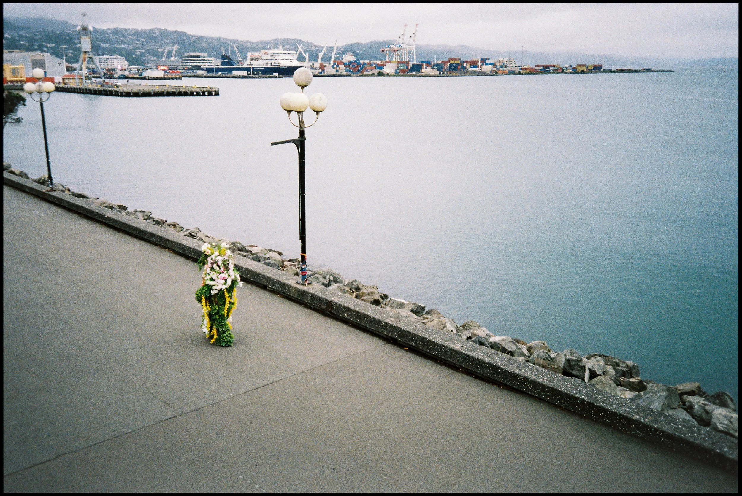 A memorial with flowers placed by a lamppost along a waterfront promenade, with ships and container cranes in the background.