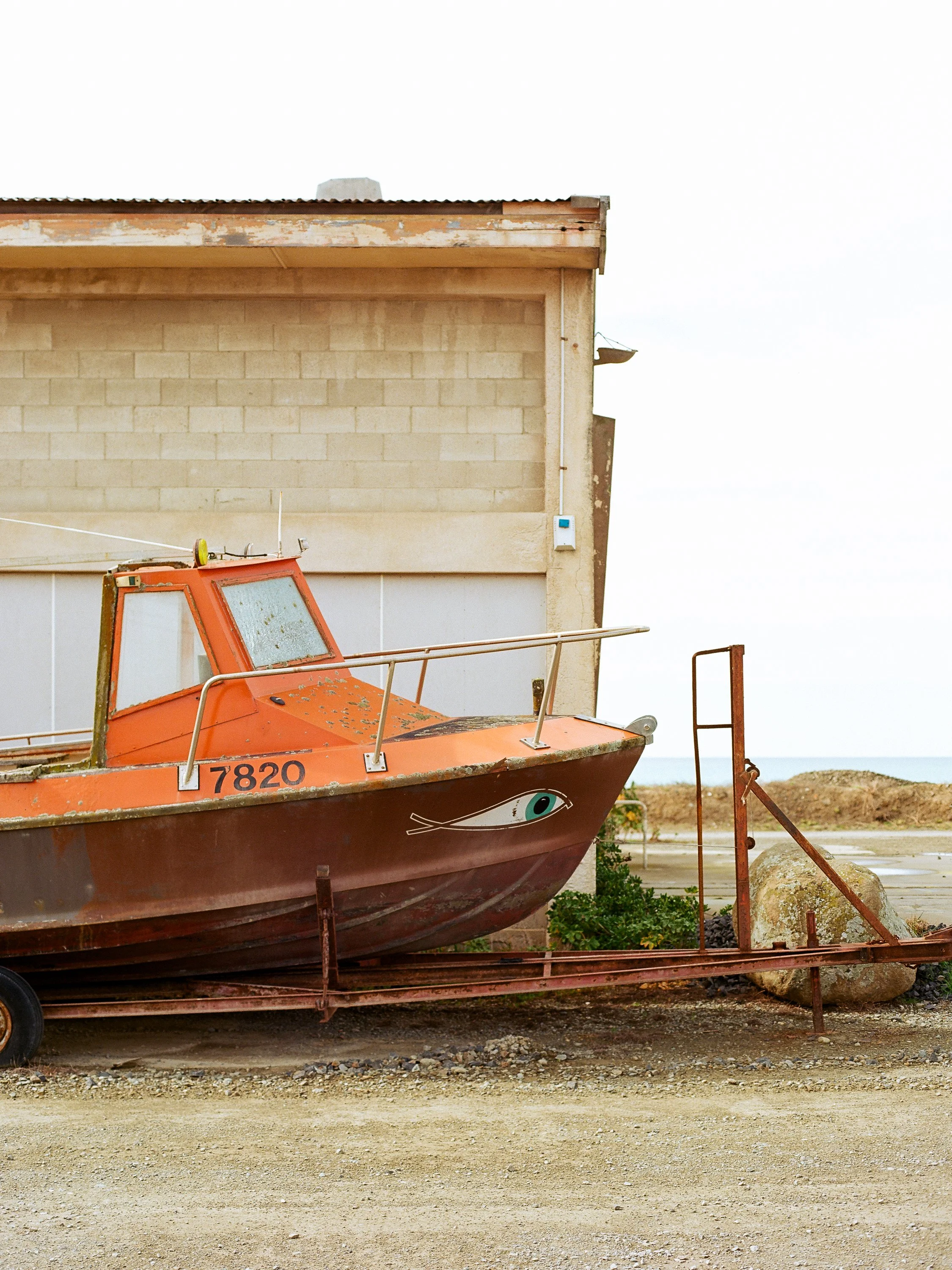 Old orange boat on a rusty trailer in front of a building near the beach.