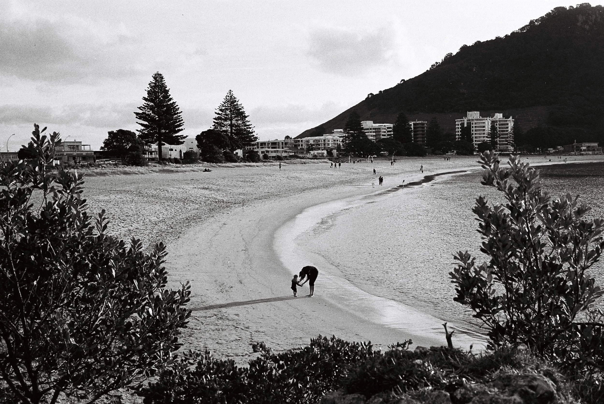 A black and white photo of a beach with people walking along the shore, including an adult and child holding hands in the foreground. There are trees and residential buildings in the background, with a hillside on the right.