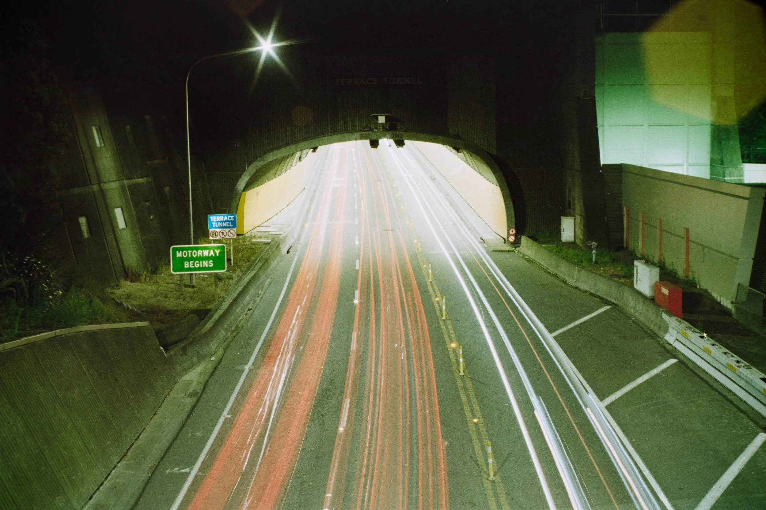 Long exposure photo of a highway passing through a tunnel at night, with light trails from passing vehicles and green signs reading "Terrace Tunnel" and "Motorway Begins".
