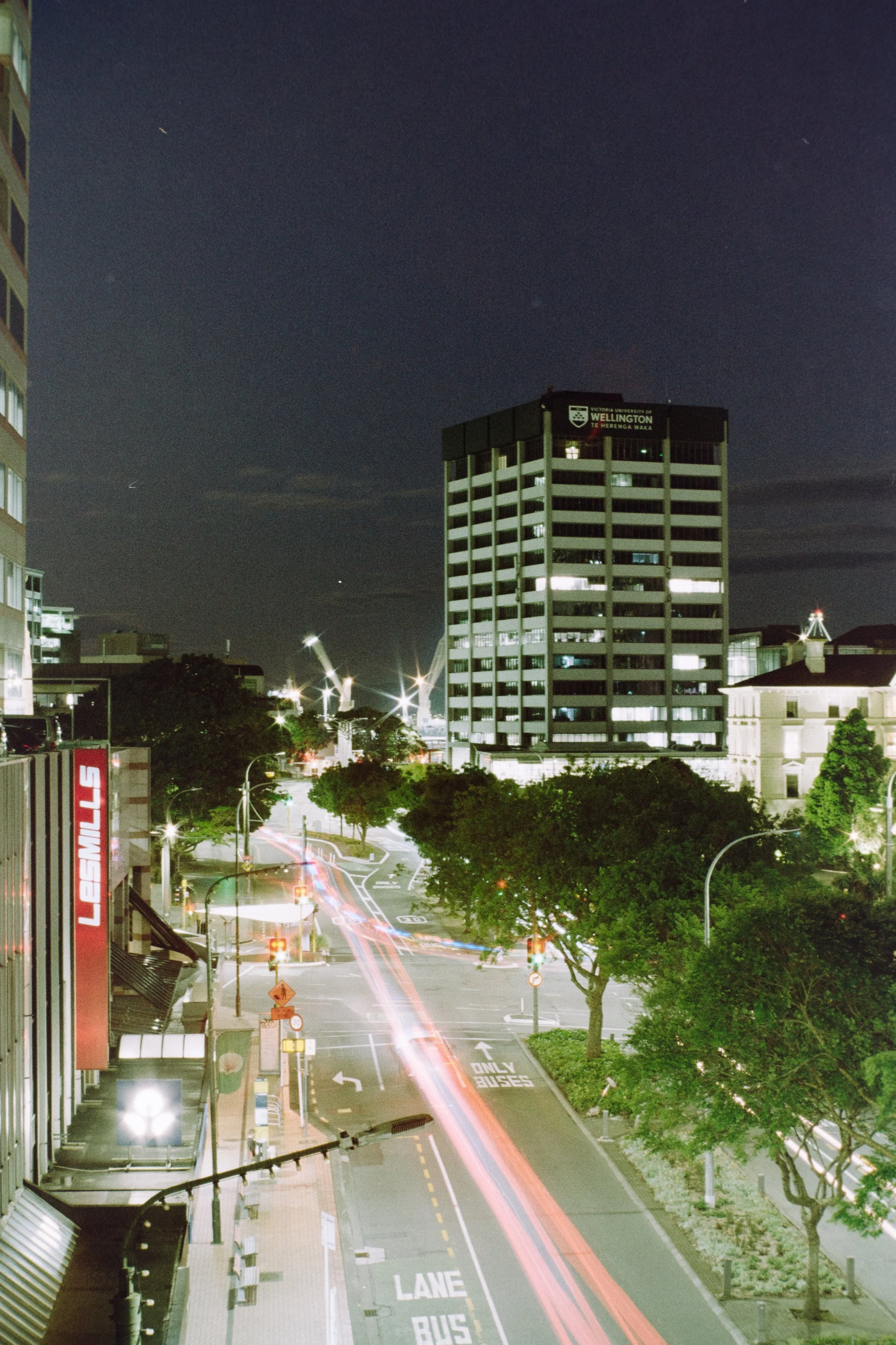 Night cityscape with tall building, trees, and streaks of light from moving vehicles.