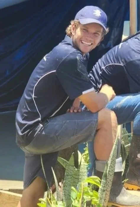 A man wearing a blue cap and dark athletic shirt, smiling, sitting outdoors near a body of water with plants in front, in a casual setting.
