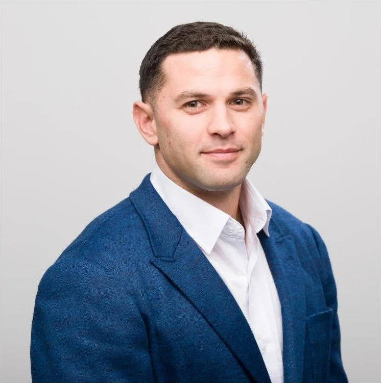A man with short dark hair wearing a white collared shirt and a blue blazer, posing against a plain light gray background.