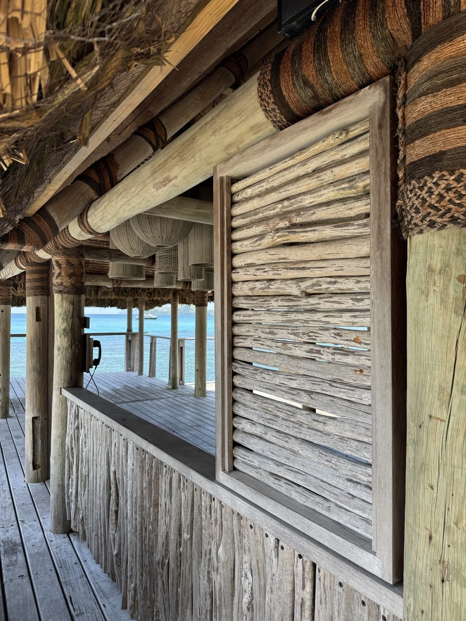 Wooden beach hut with straw roof and bamboo walls, overlooking the ocean with a boat in the water.